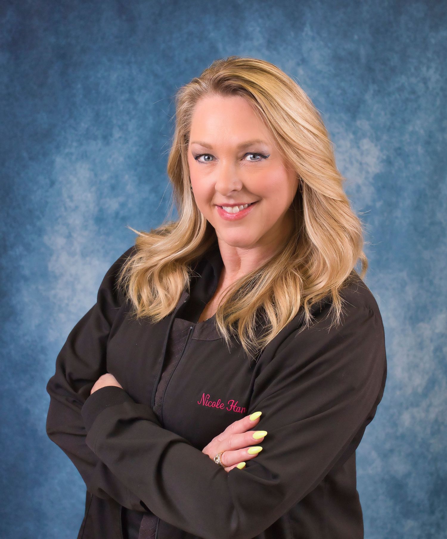 Smiling blonde woman in dark embroidered jacket posing professionally against blue studio background.