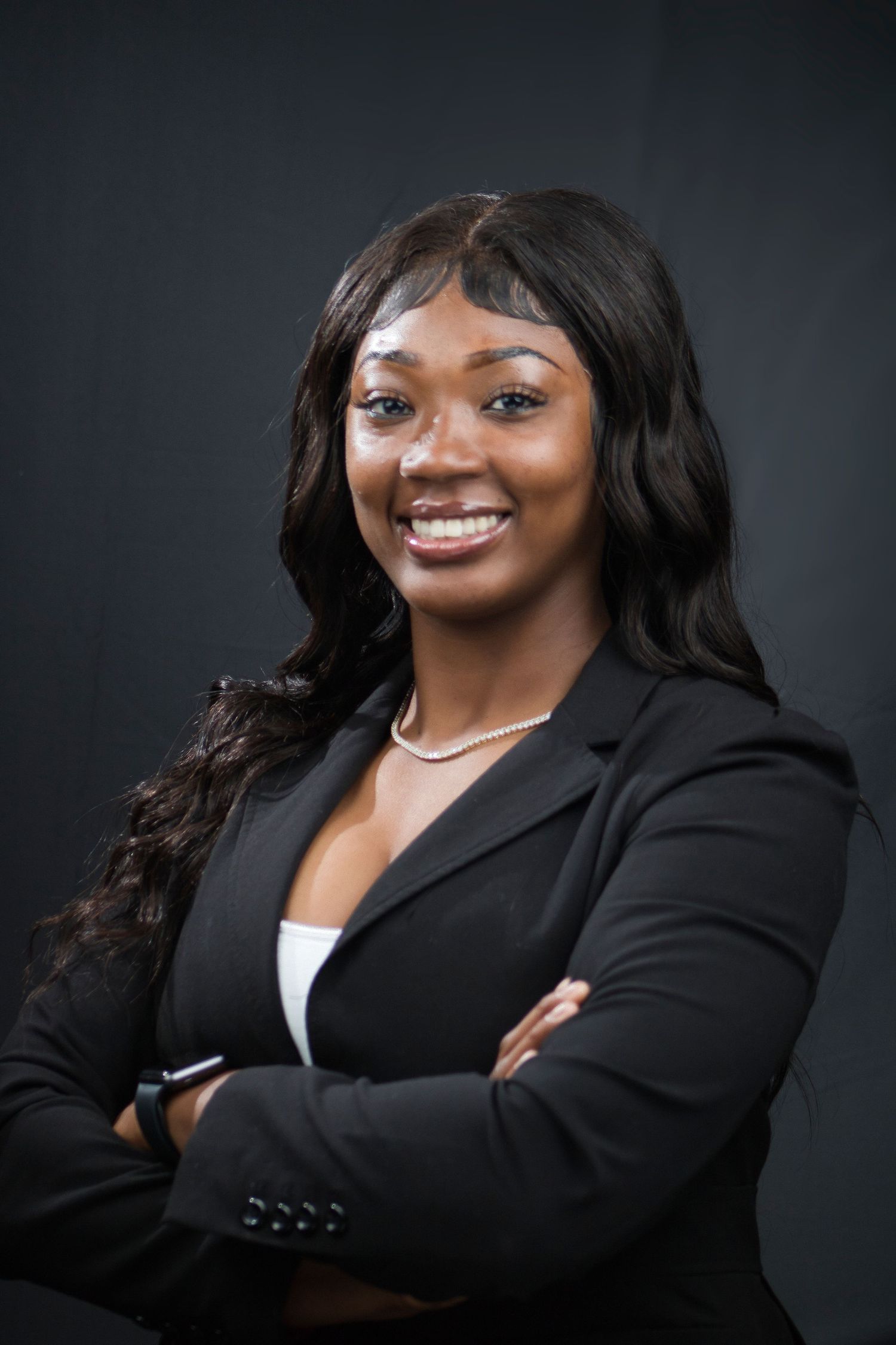 Professional Black woman in black blazer smiling confidently with arms crossed against dark background.