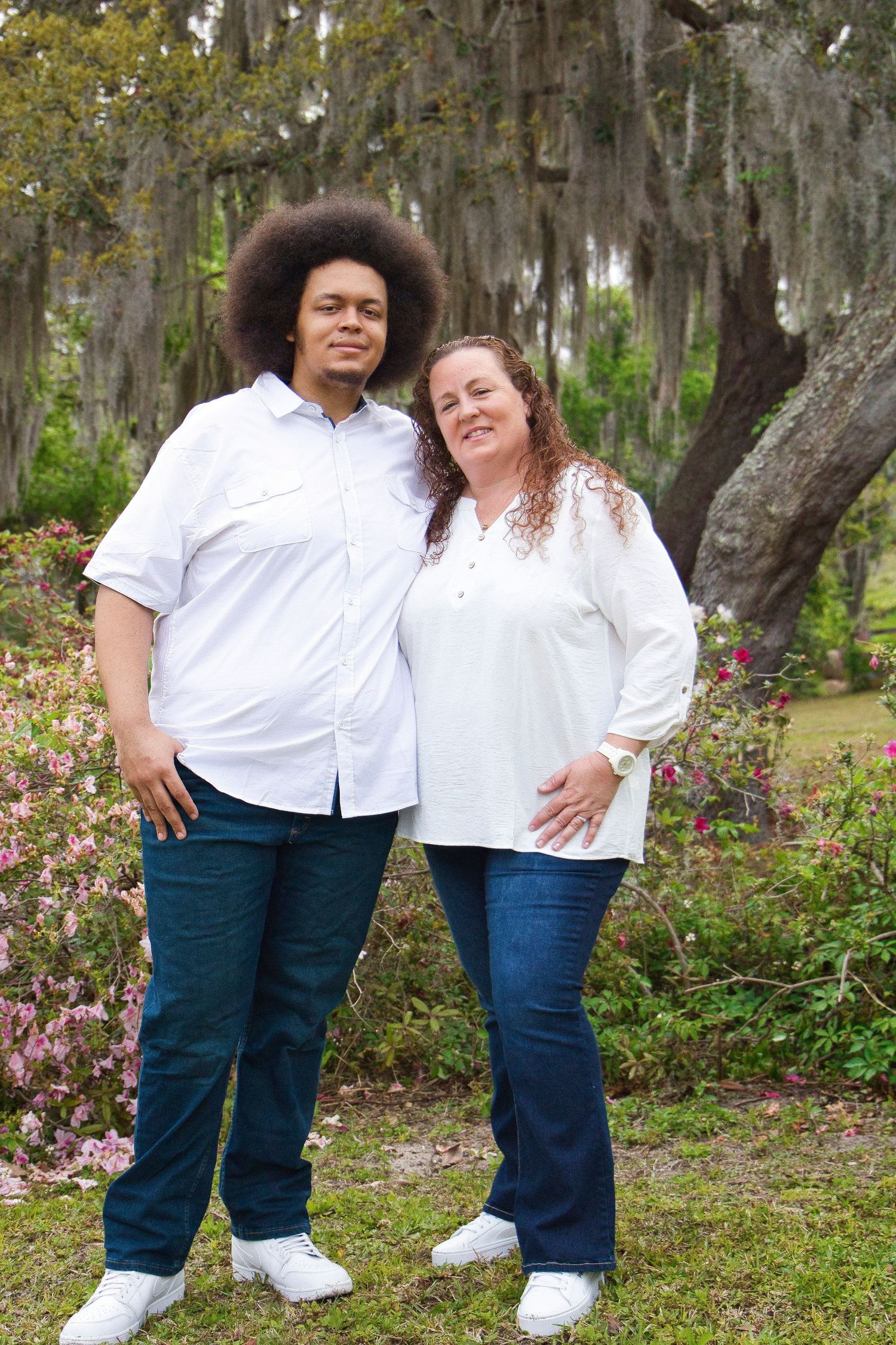 Interracial couple in matching white shirts and jeans smiling outdoors near blooming azaleas and Spanish moss-draped oak tree.