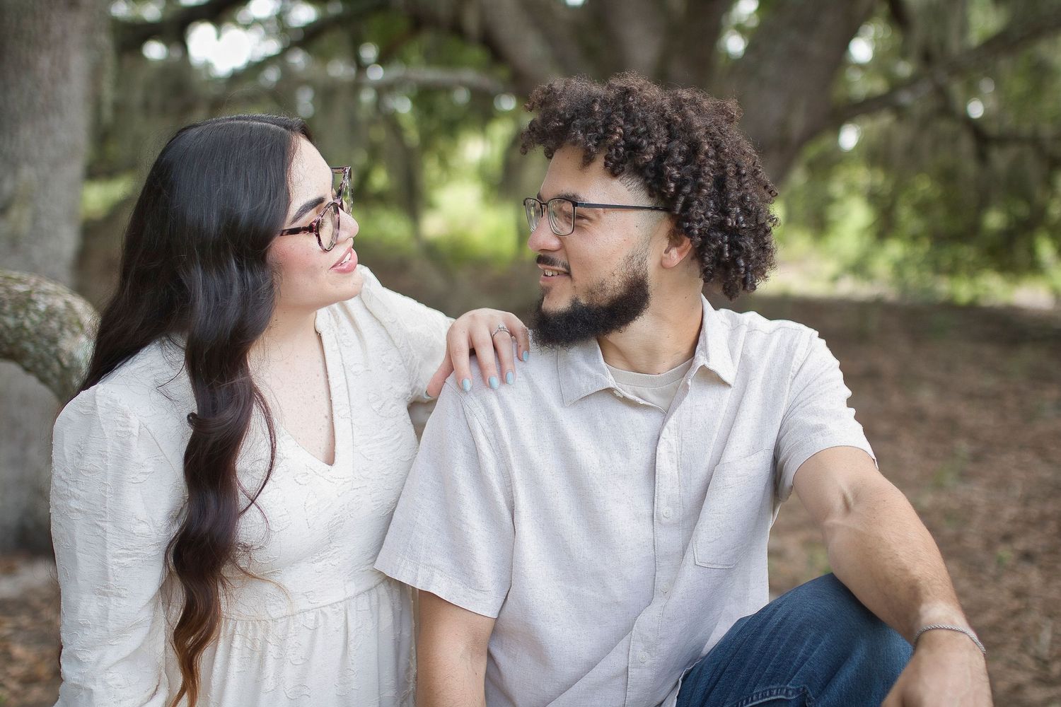 A young couple sits together outdoors under a tree, smiling at each other in a romantic portrait setting.