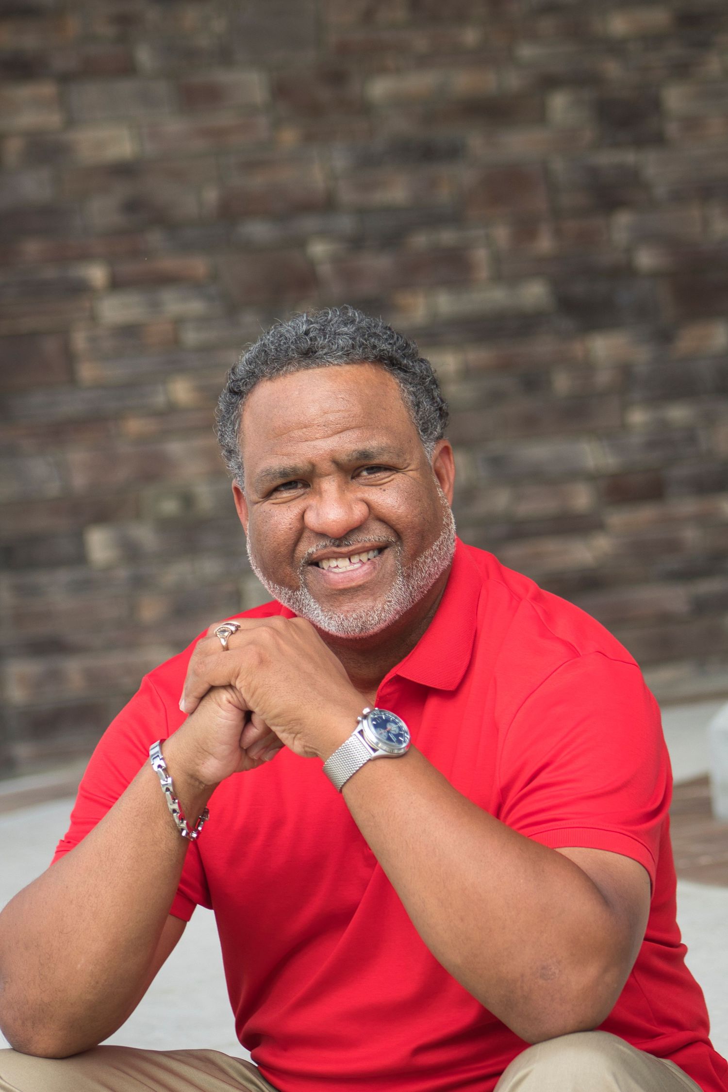 Smiling middle-aged Black man in red polo shirt sitting outdoors against brick wall with hands clasped, wearing a watch.