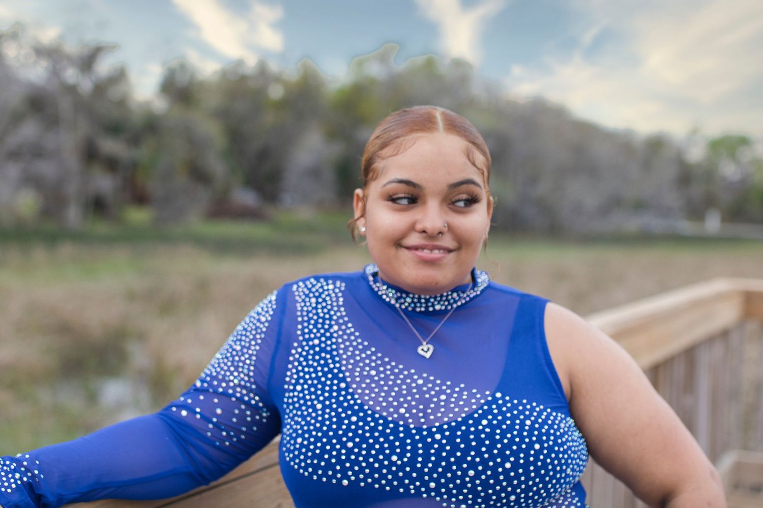 A young woman in a blue rhinestone bodysuit smiles on a wooden bridge with a scenic outdoor backdrop.