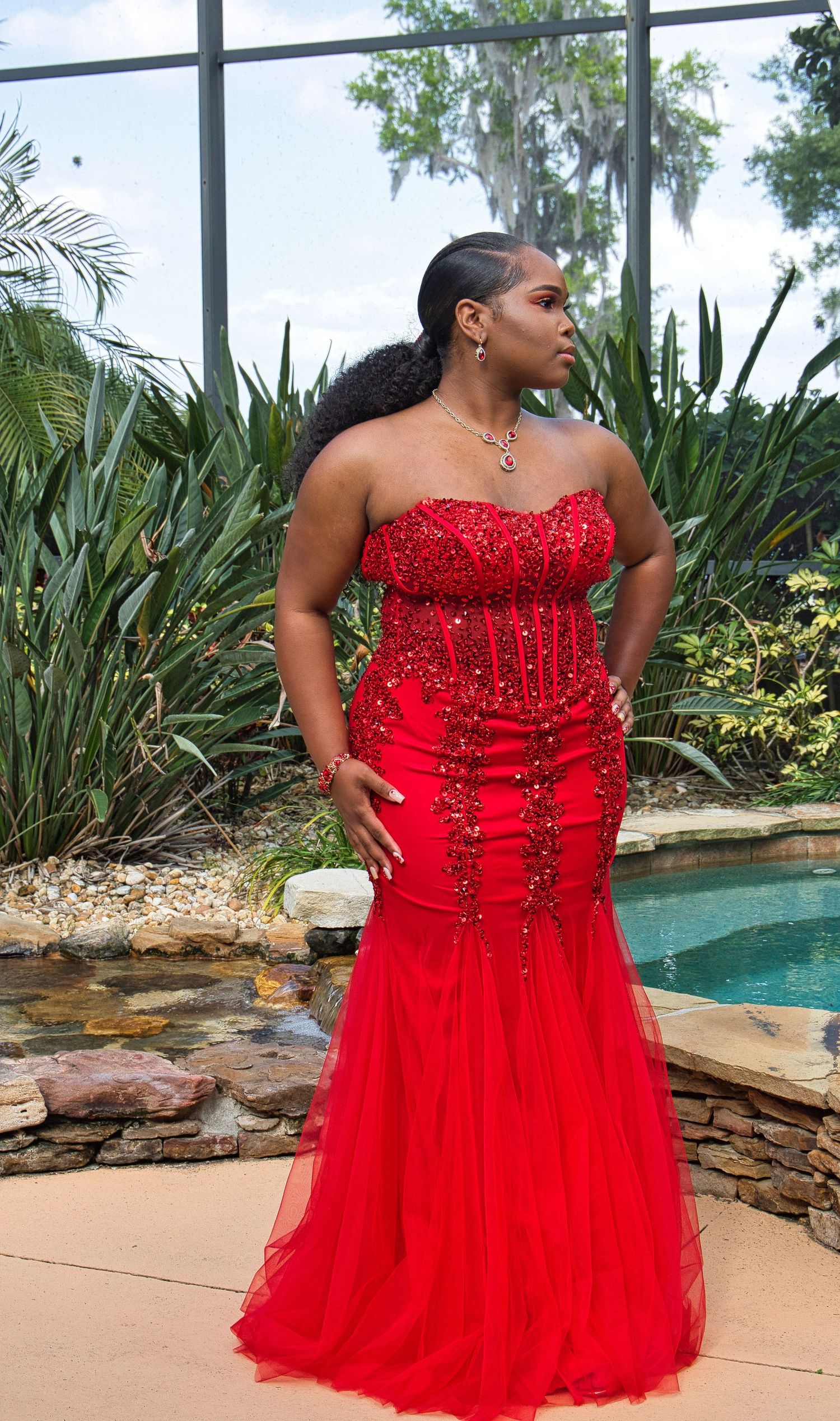 Glamorous red beaded evening dress with sweetheart neckline shown against tropical landscaping.