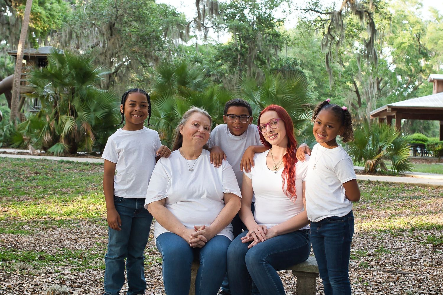 Family of five smiling together outdoors in a park, wearing matching white shirts and jeans.