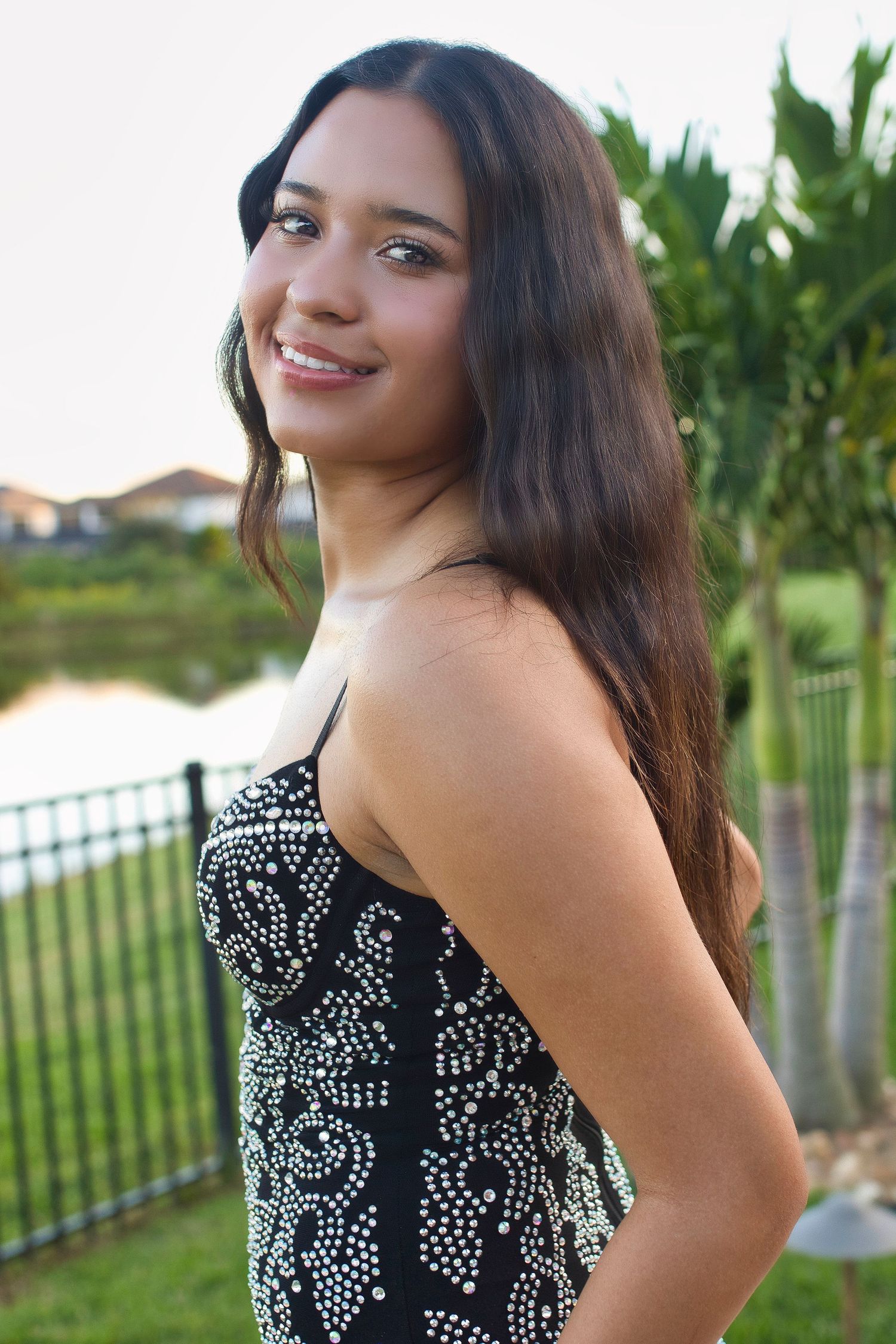 Young woman in black beaded dress smiling outdoors near a lake with lush greenery and fence in background.