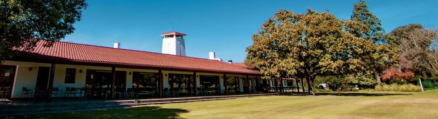 Long single-story building with a red metal roof and covered veranda beside a green lawn, shaded by mature trees under a clear blue sky.