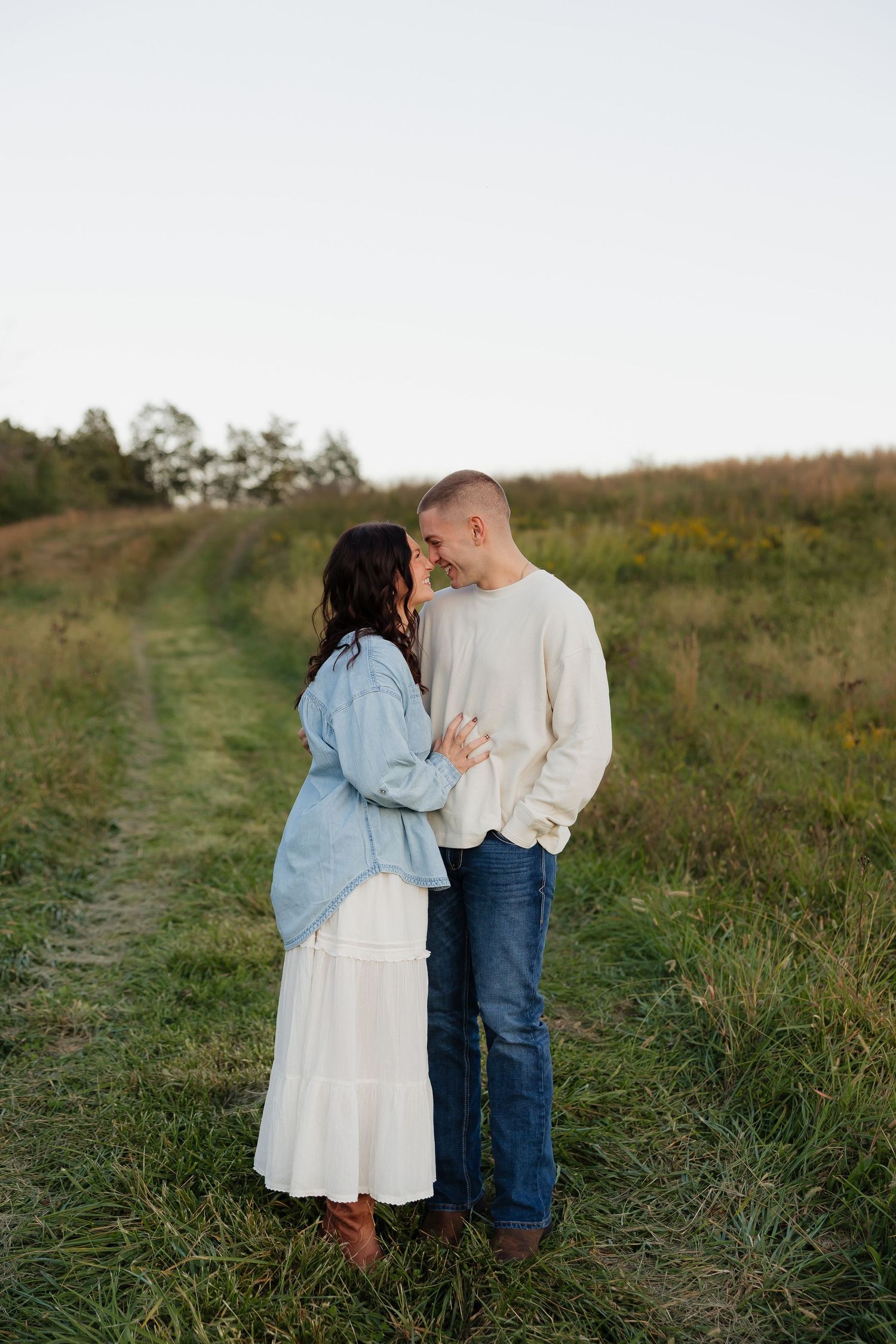 Couple sharing a romantic moment on a grassy field path, woman in white skirt and denim jacket, man in cream sweater and jeans.