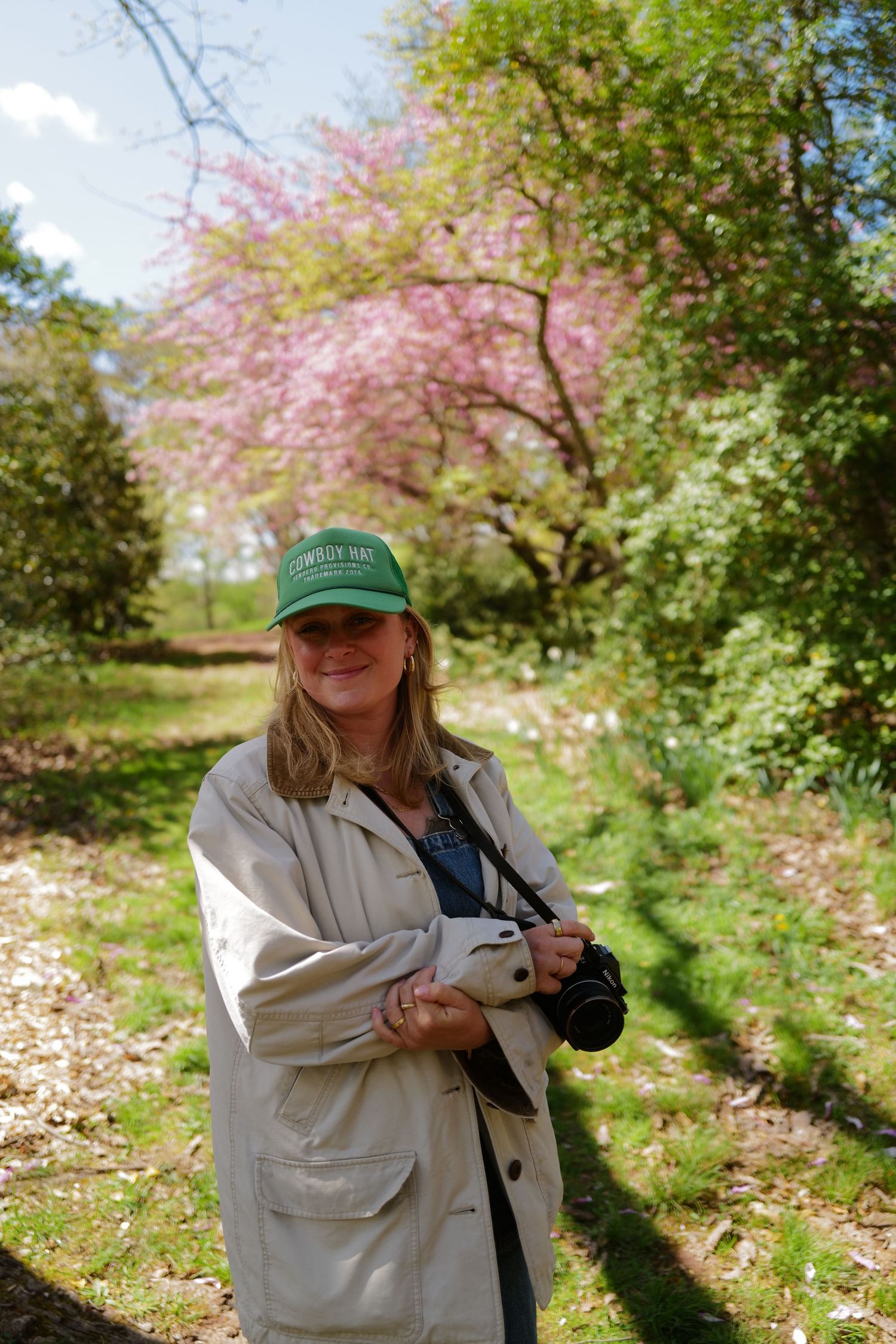 A photographer holding a camera stands on a path beneath blooming pink cherry blossom trees in springtime.