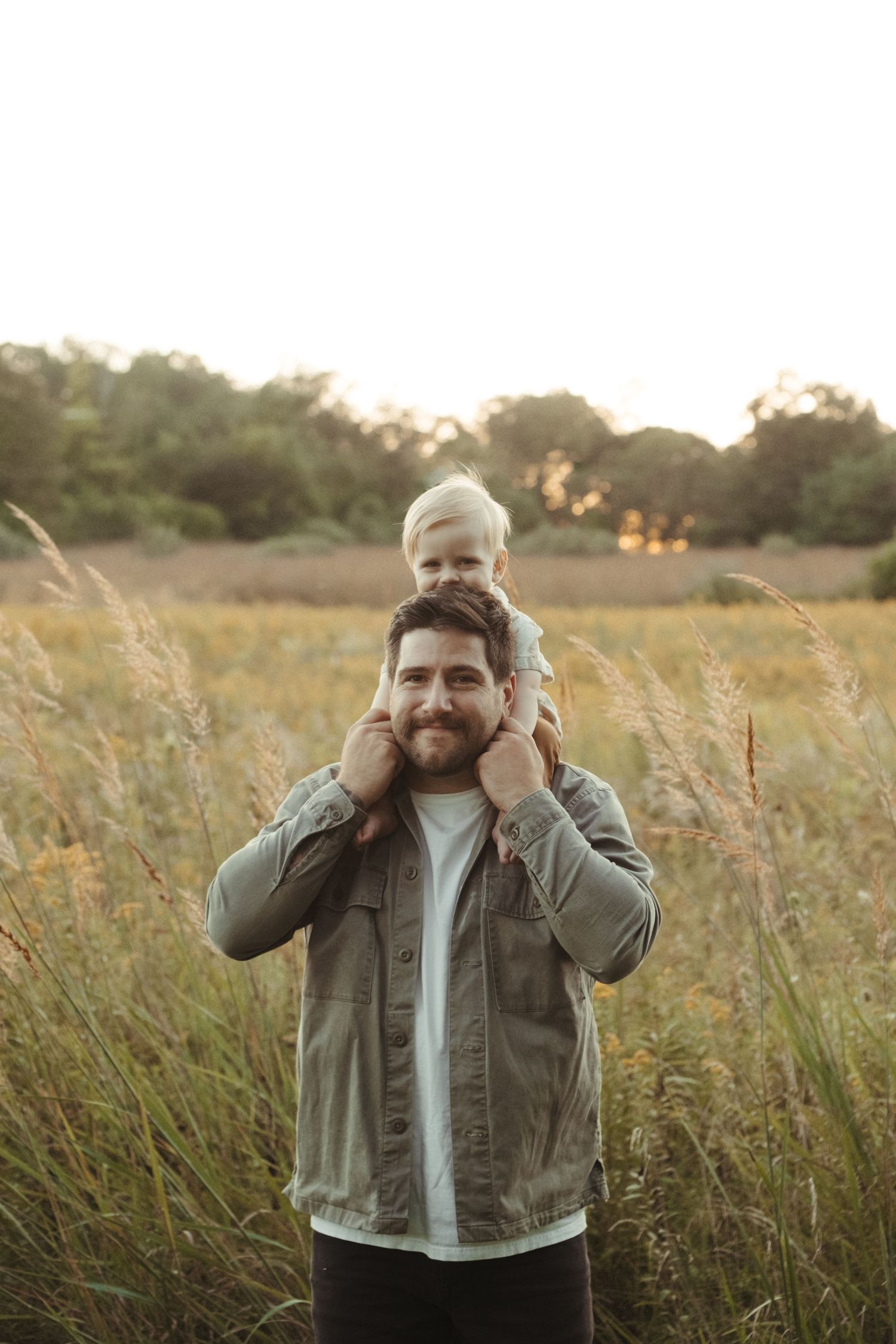 A toddler sits on shoulders in a golden field at sunset while tall grasses sway in the warm evening light in califon nj.