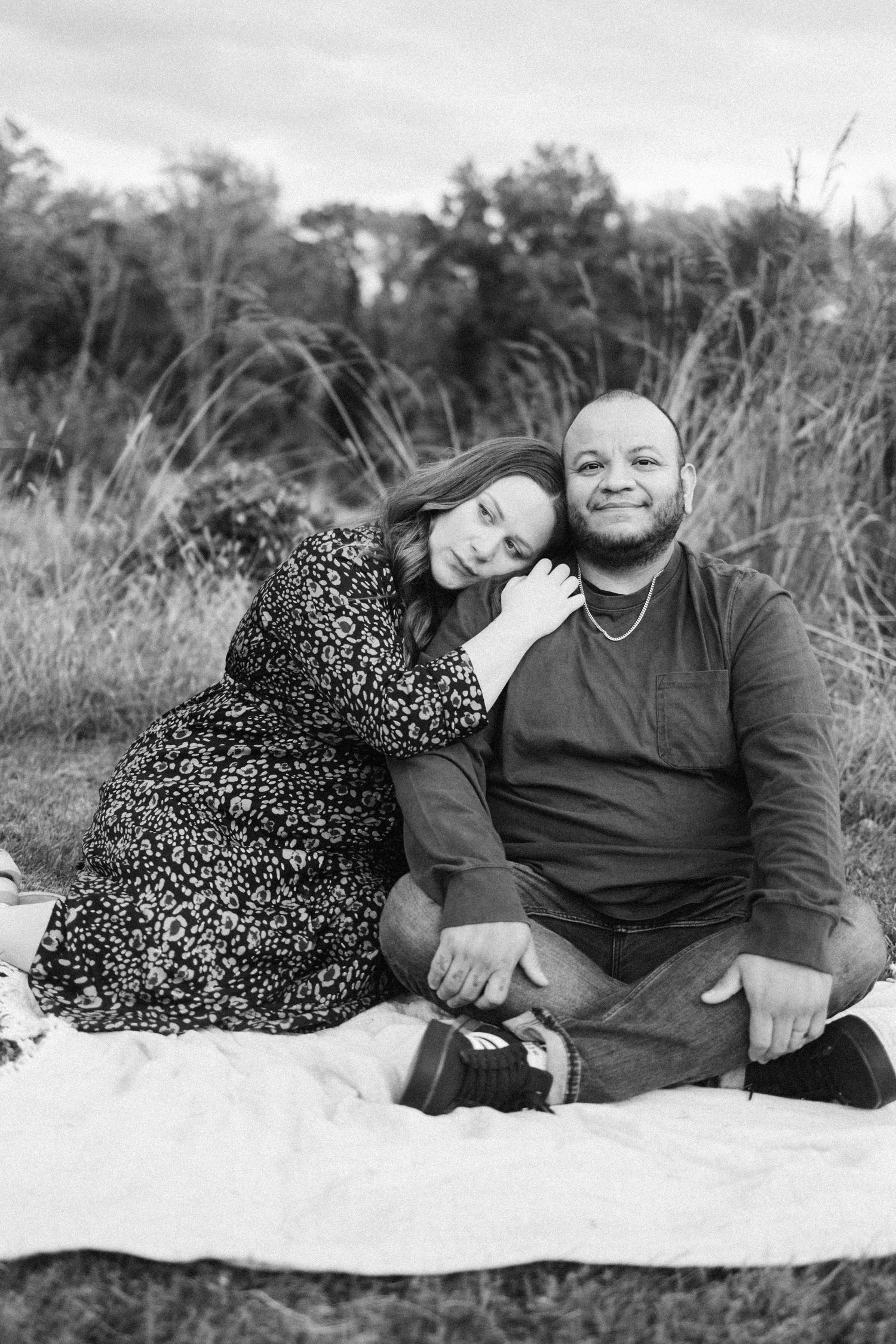 A couple sits together on a blanket outdoors in a black and white photo featuring tall grass and nature in the background in hunterdon nj.