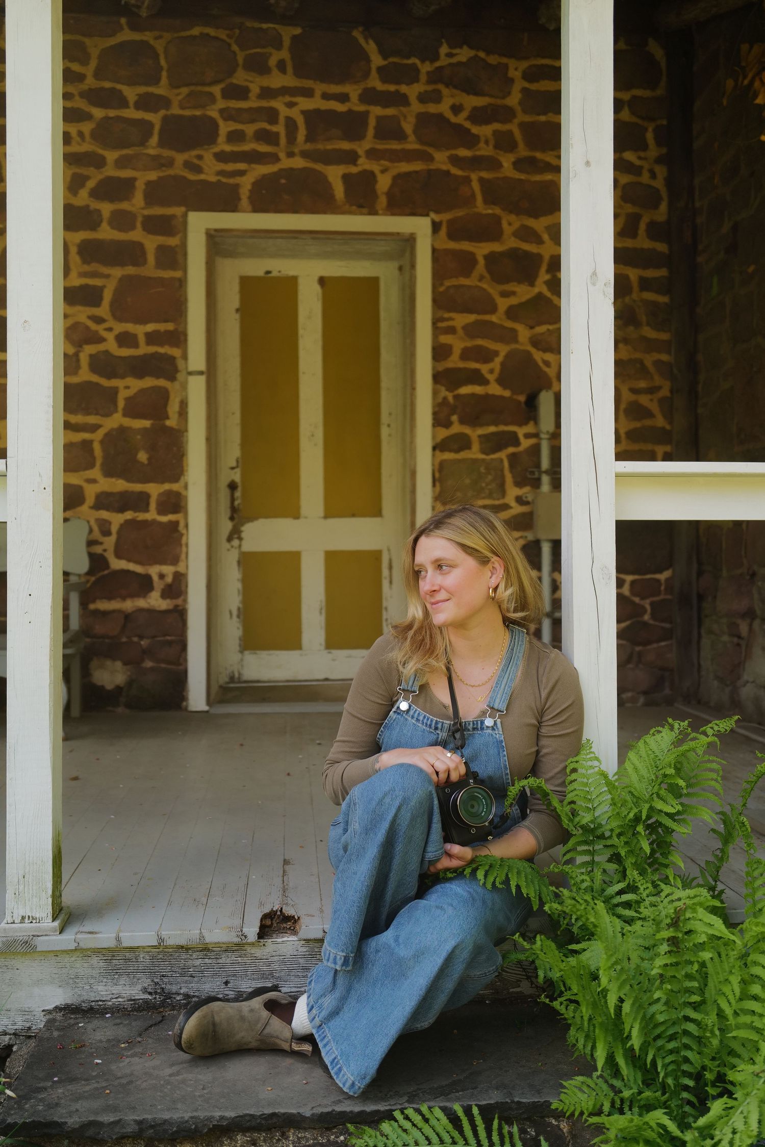 Someone sits in overalls on a rustic stone house porch with wooden columns and ferns growing nearby.
