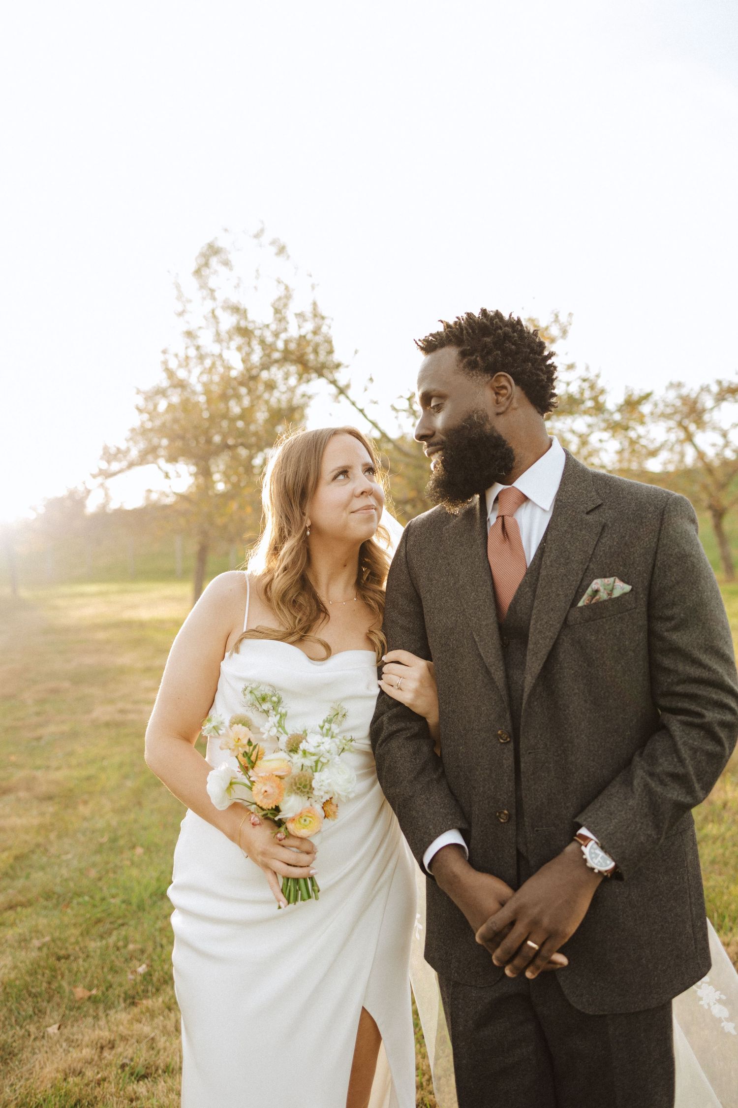 A couple in wedding attire stand together in a sun-drenched field during golden hour, the bride holding a bouquet in vernon nj.