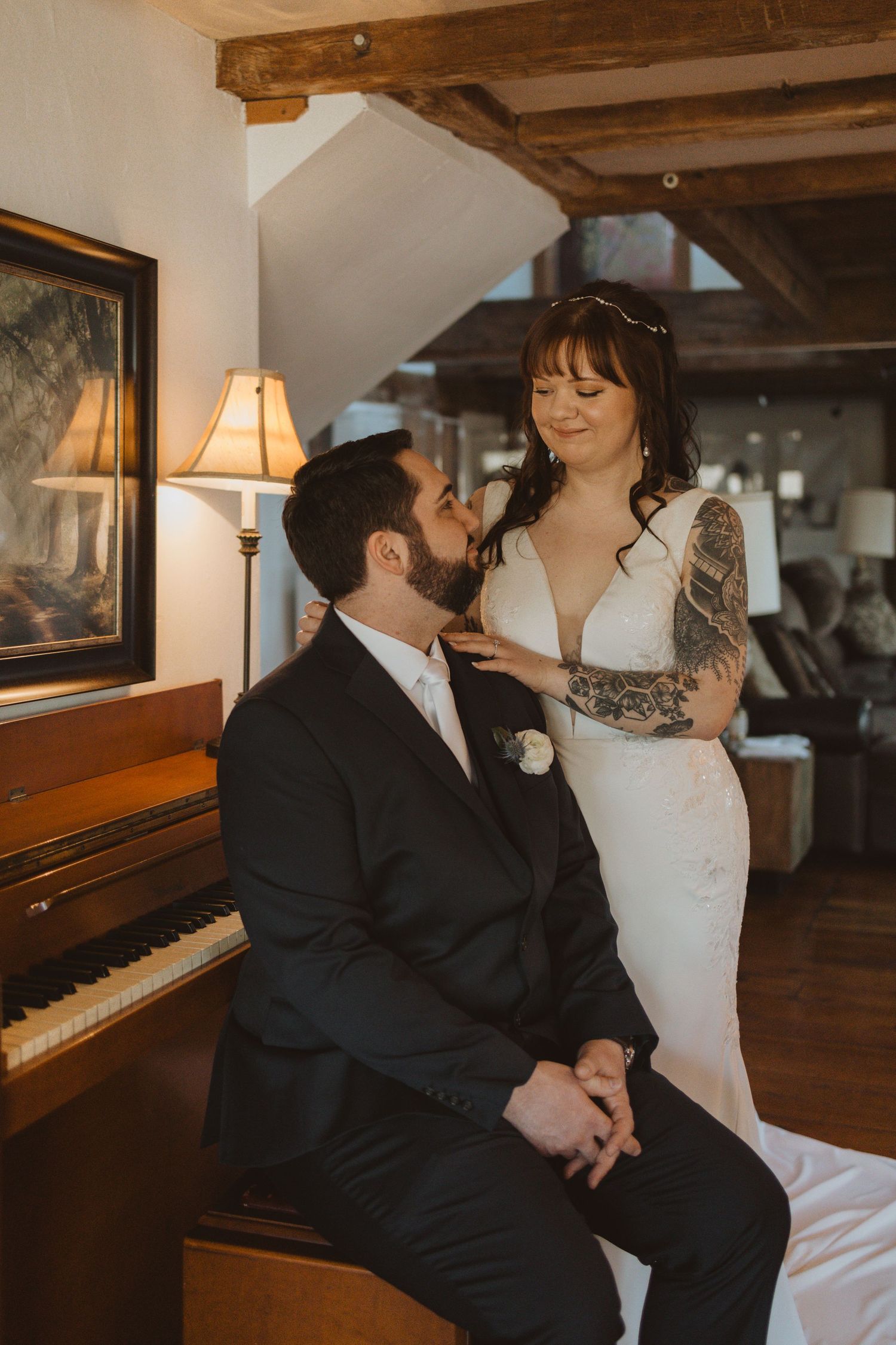 Intimate wedding moment captured near piano with bride in sleek white dress and groom in dark suit in rustic setting.