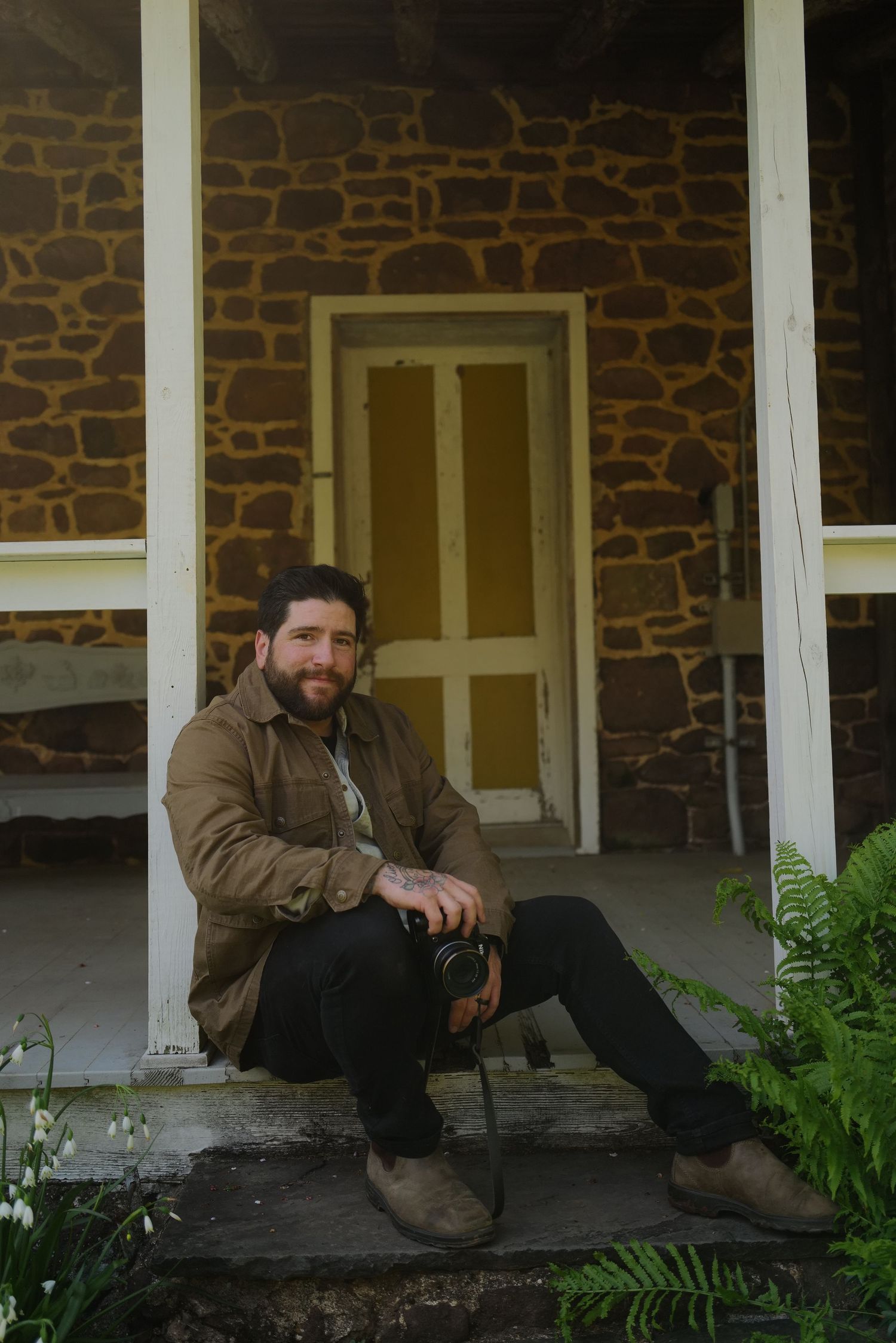 Someone sits on stone porch steps of a rustic building with stone walls and a wooden door, surrounded by ferns.