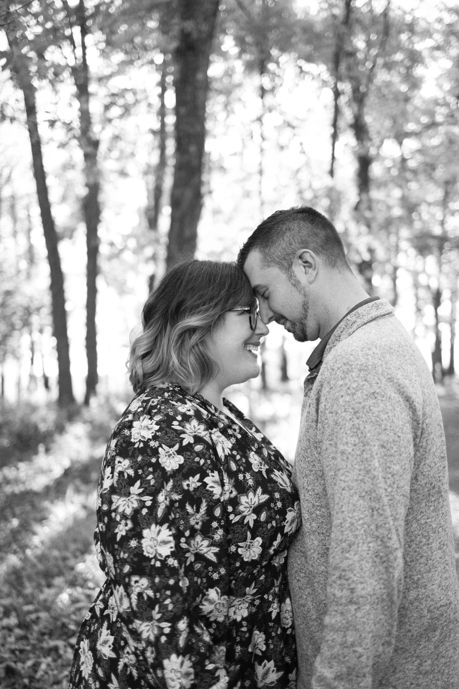 A loving couple shares an intimate forehead touch moment in a sunlit forest setting, with a floral dress and sweater in hunterdon county.