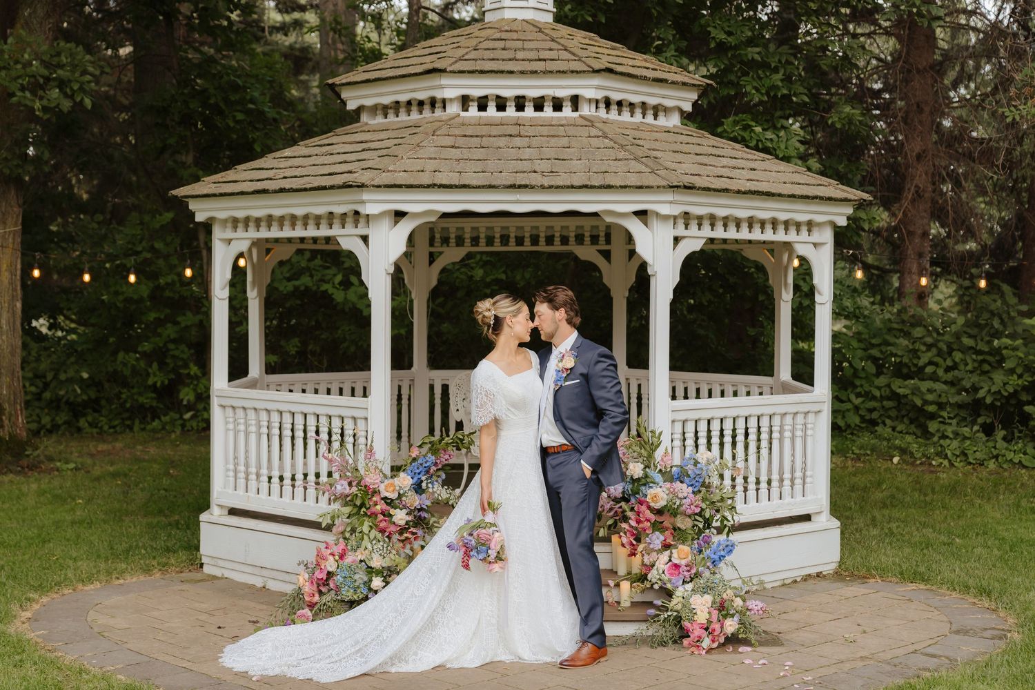 A wedding couple posing in front of a white gazebo decorated with pink floral arrangements.