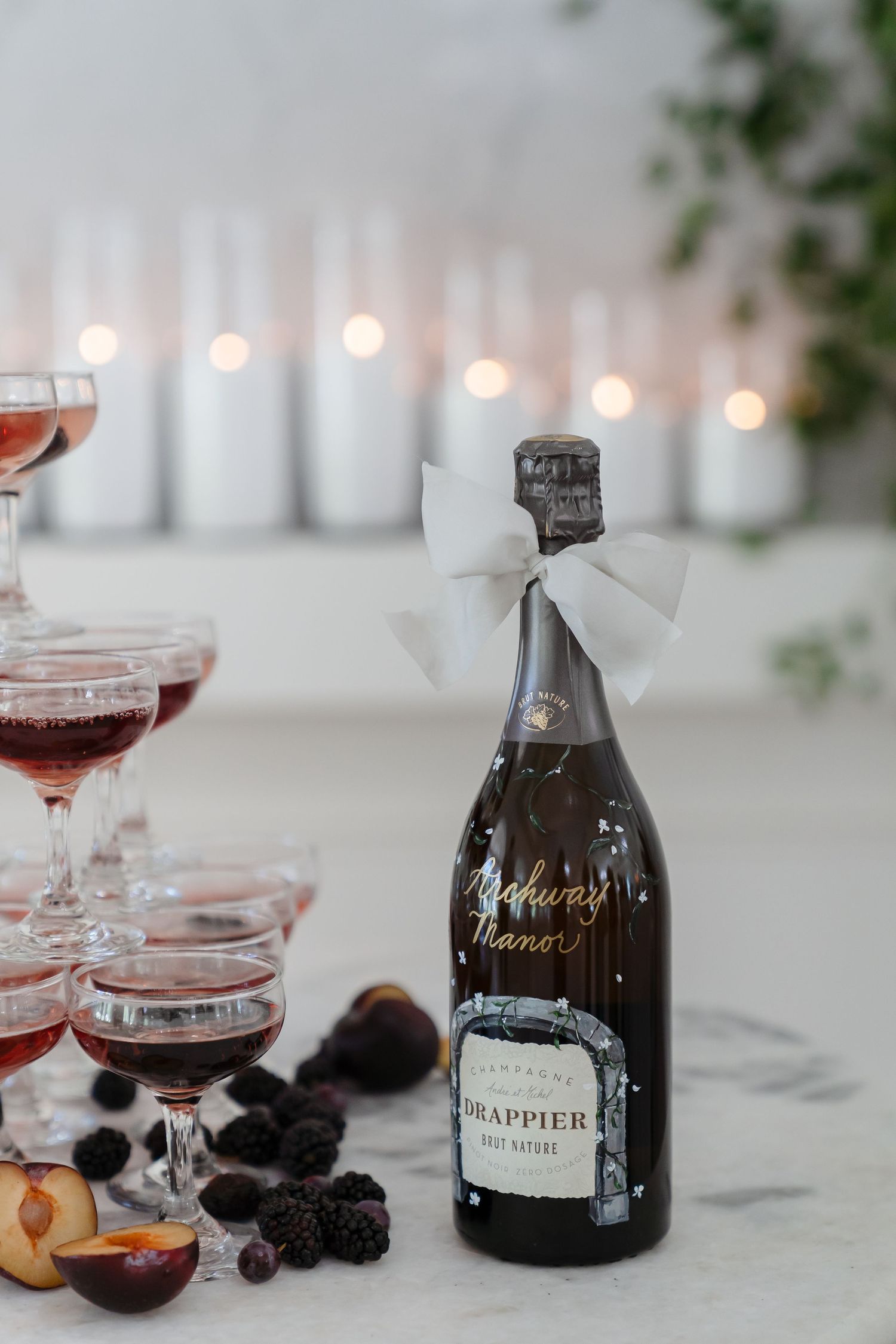 Elegant champagne bottle adorned with white bow next to wine glasses and fresh fruit on white tablecloth.