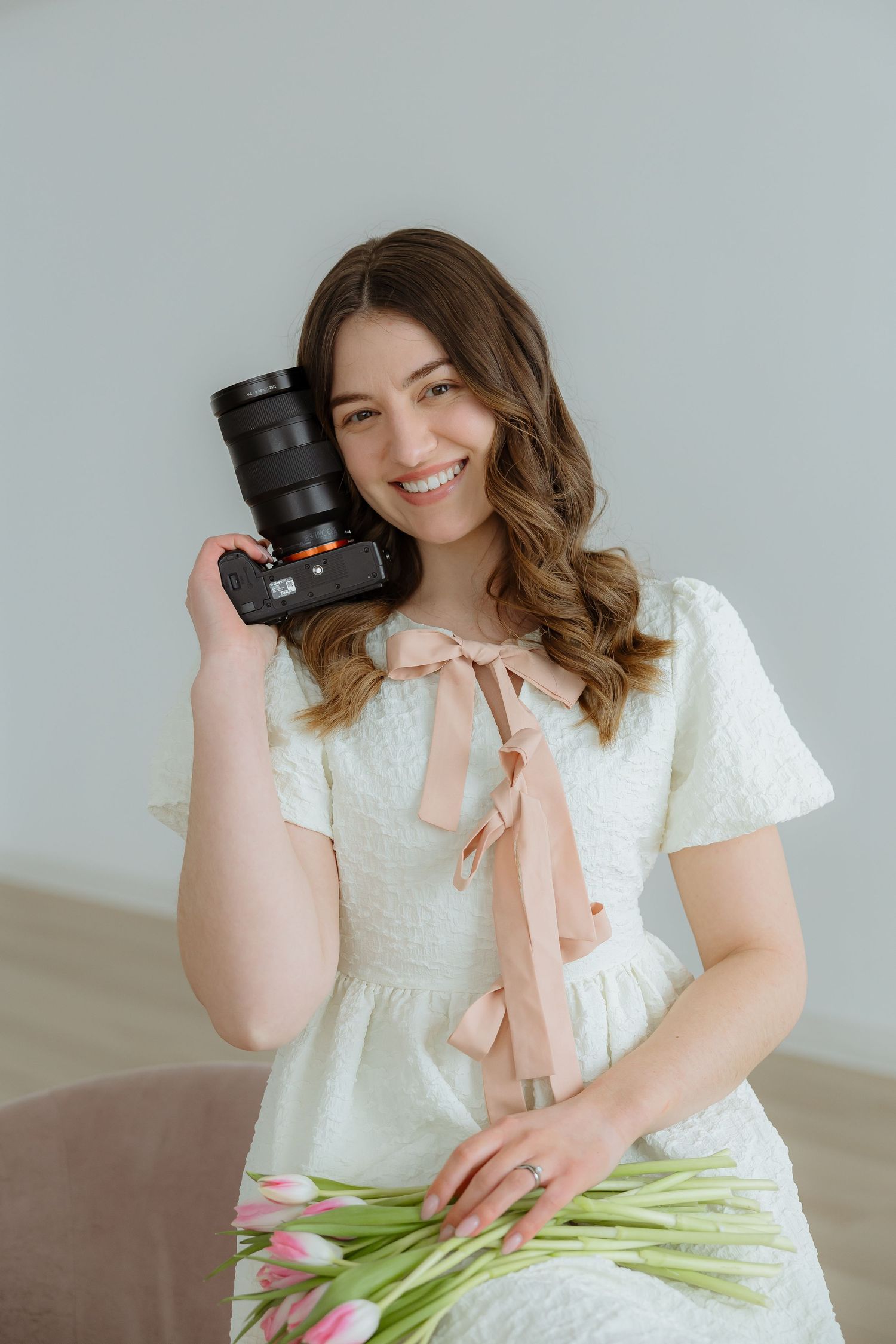 Sara Wiebe a pofessional couples and wedding photographer in white dress holds a camera and tulips against gray background.