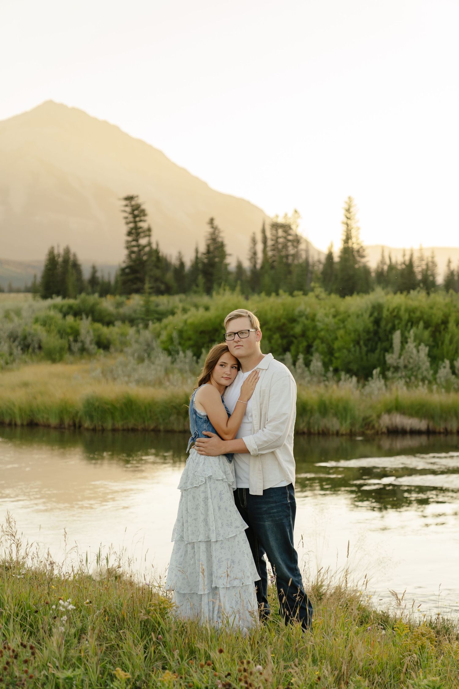 A couple shares an intimate moment by a serene lake at sunset with mountains and evergreen trees in the background.