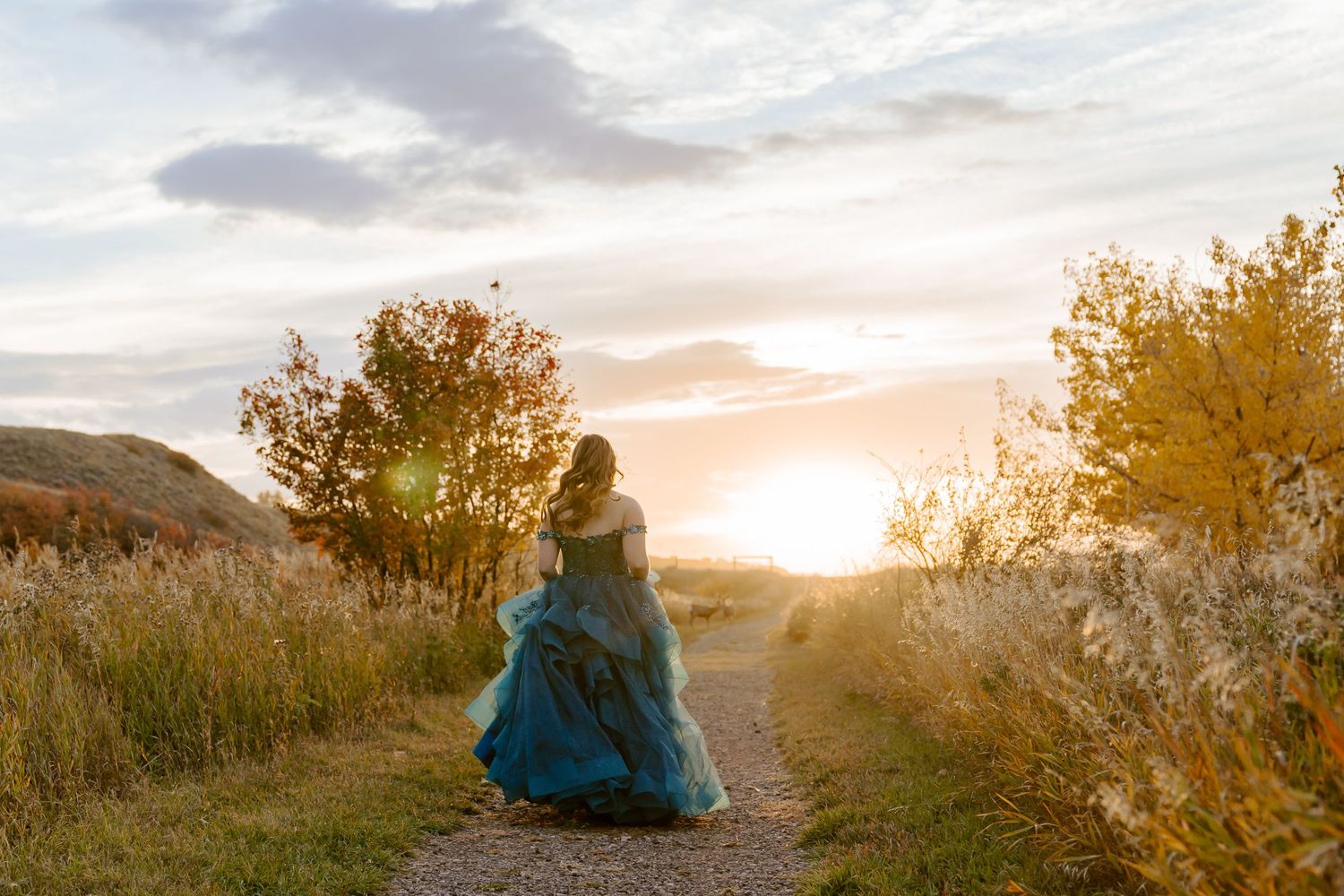 Silhouette in a flowing blue dress walks down a path at sunset with autumn trees in the background.