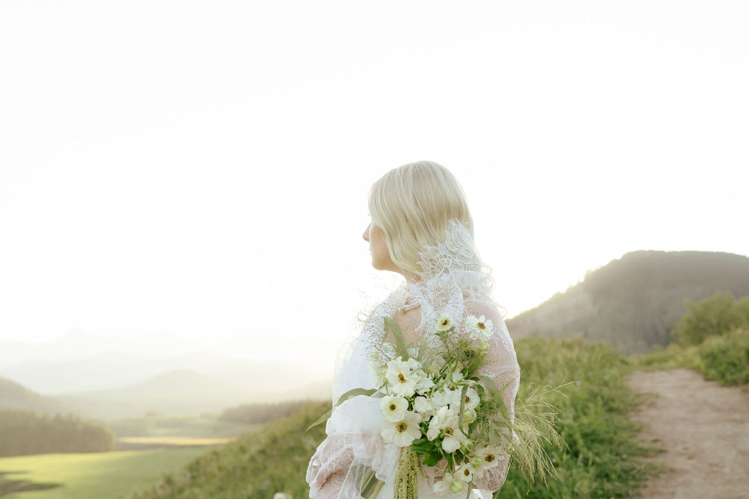 A wedding couple stands on a scenic coastal cliff at sunset with the ocean and mountains in the background.