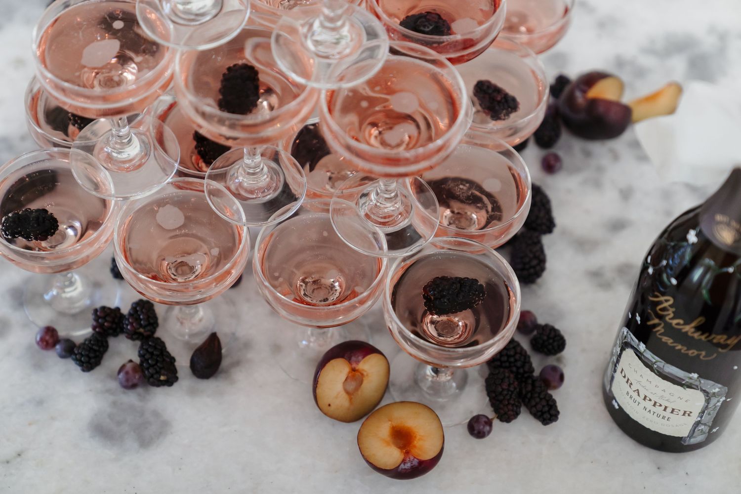 Rose-colored champagne glasses garnished with fresh fruits on marble surface.