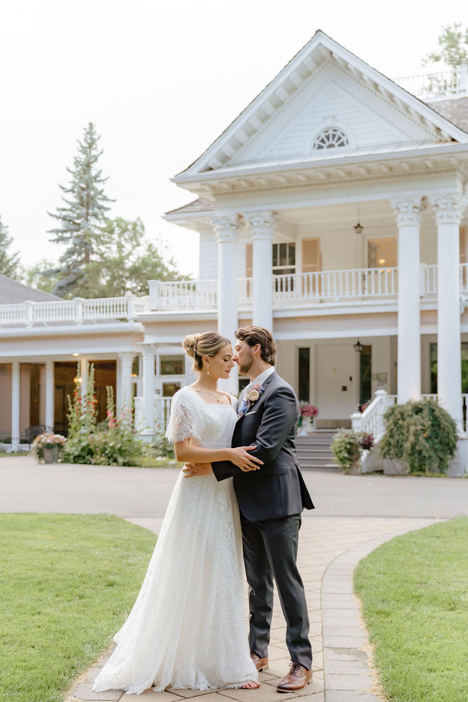 A couple shares a romantic moment outside a grand white colonial-style wedding venue with elegant columns and porches.