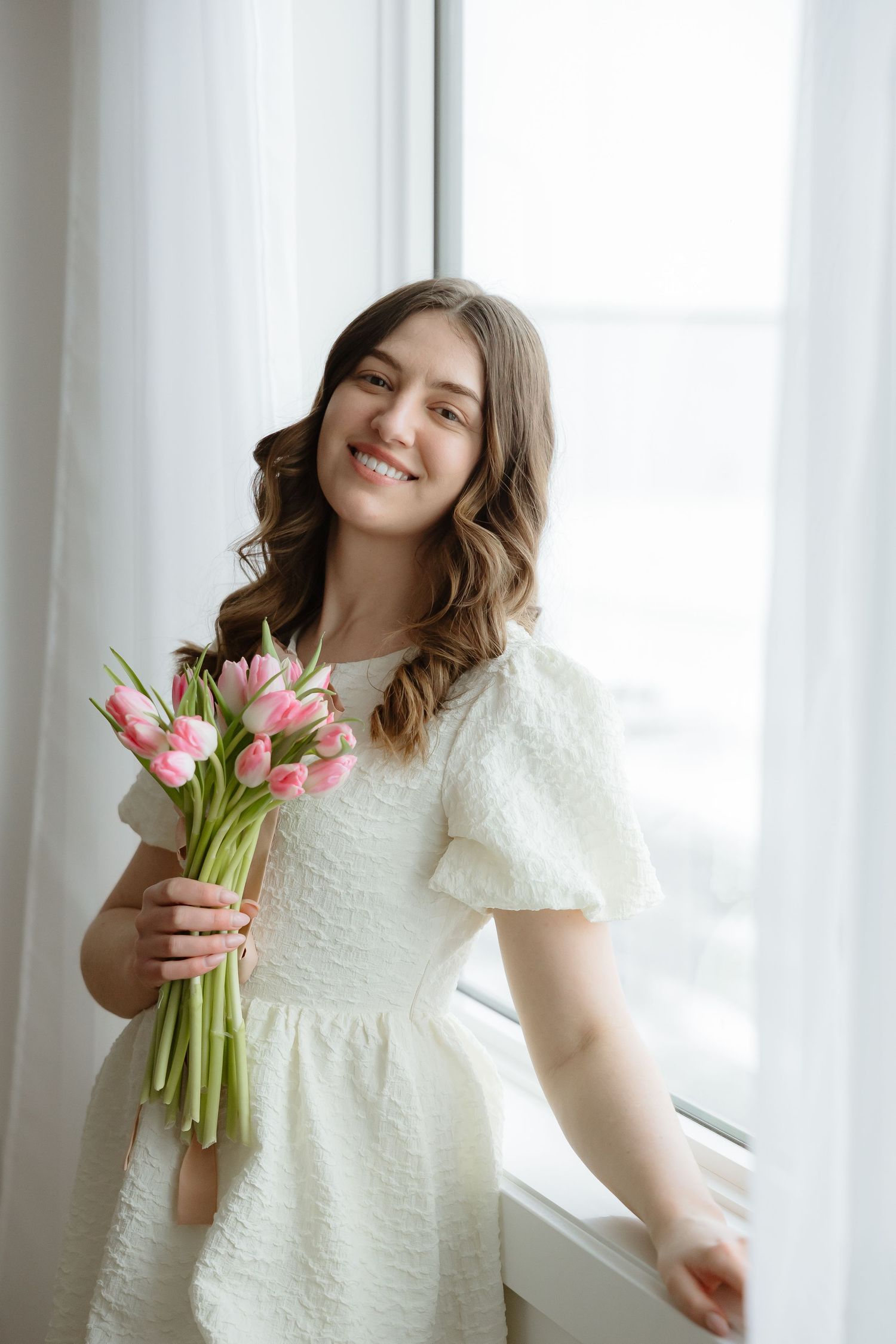 Sara Wiebe in a white dress poses with pink tulips near a sunlit window.