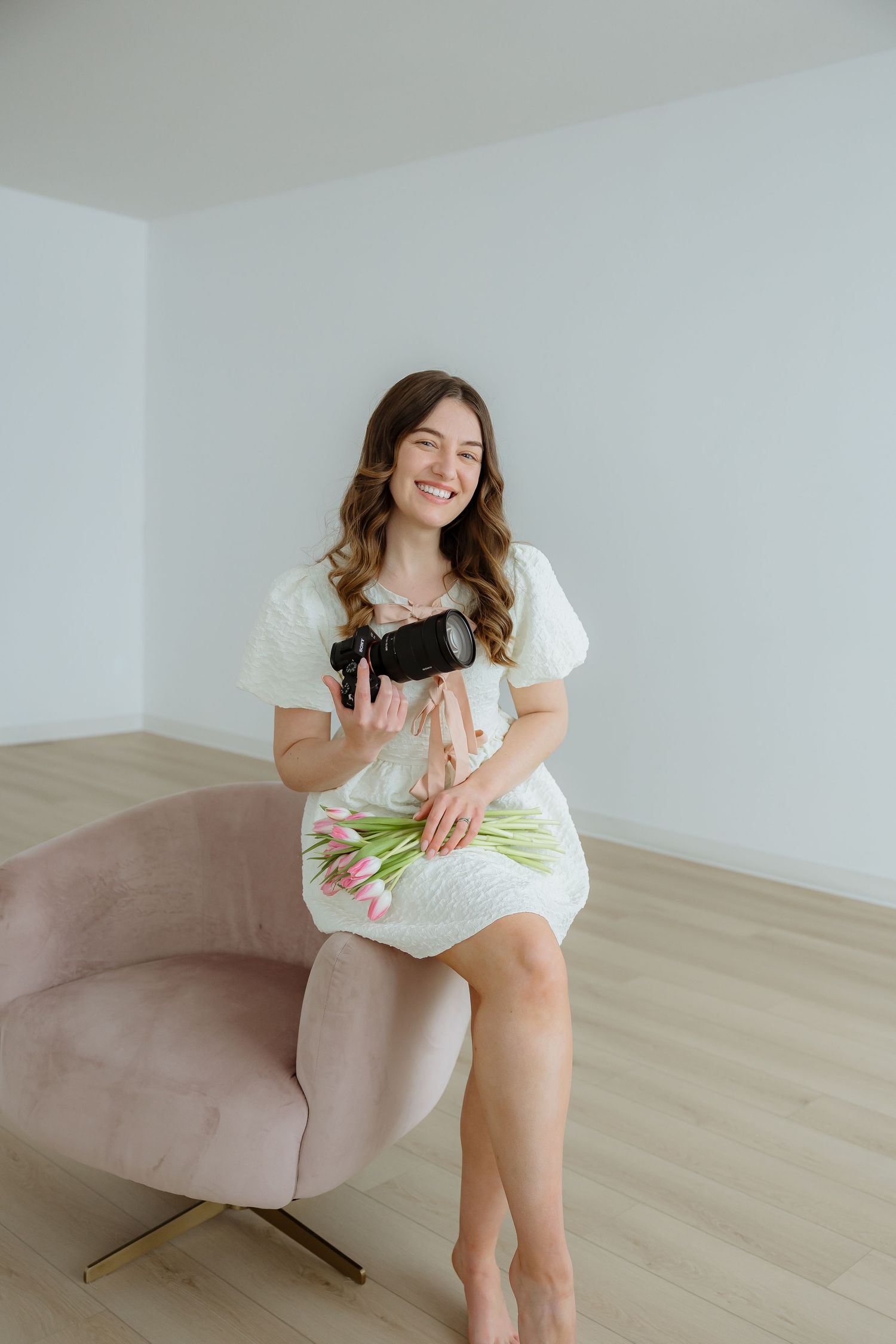 Sara Wiebe a professional photographer in white dress sitting on pink chair with flowers.
