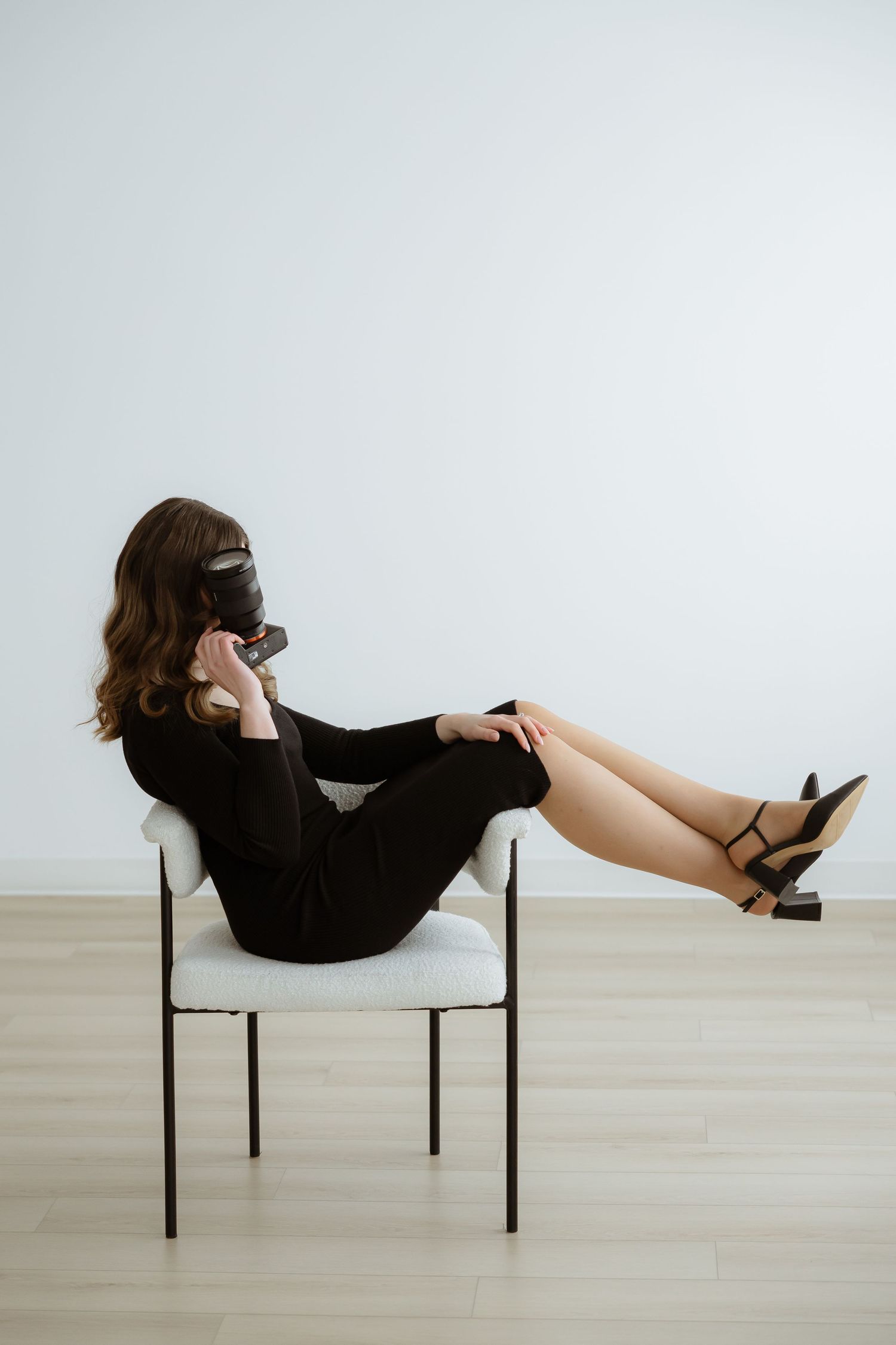 Stylish model in black dress posing on white chair against minimalist background.