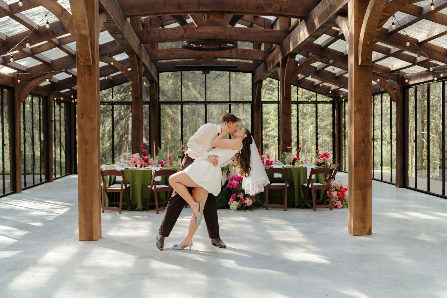 A couple performs a romantic dip dance move in a rustic glass-walled venue with wooden beams and pink floral decor.
