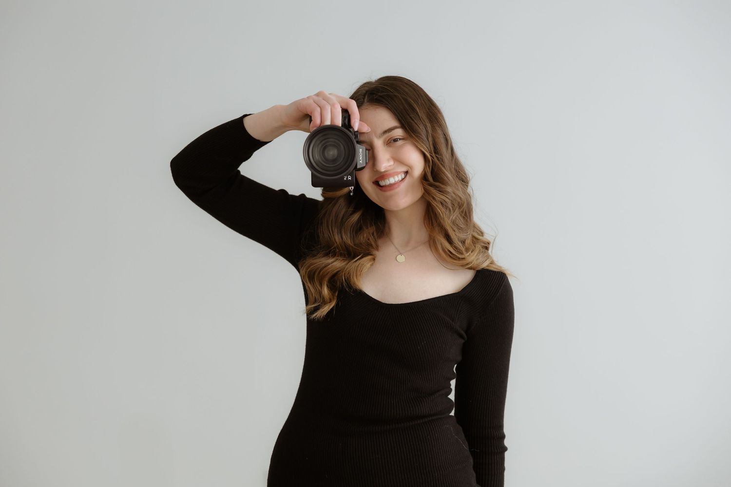 Sara Wiebe a professional couples and wedding photographer in a black sweater holding a DSLR camera against a white background.