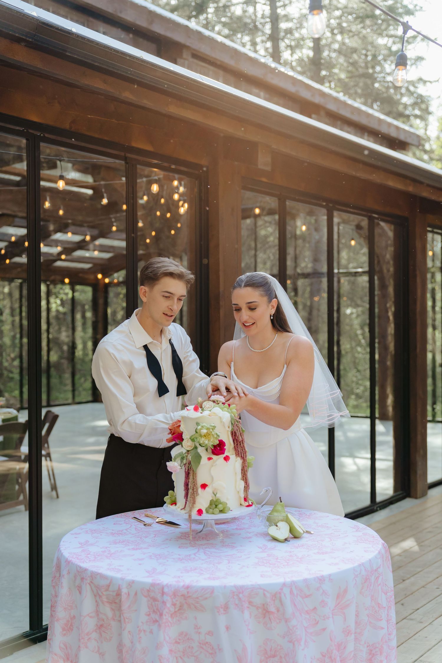 A couple in wedding attire cuts their white and pink wedding cake on an outdoor patio surrounded by trees at the Valley Weddings Venue