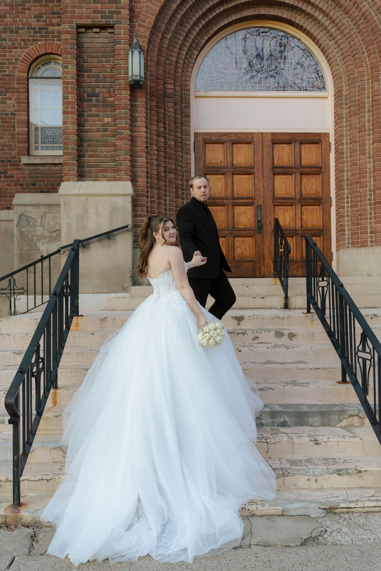 Bride in white ballgown and groom in black suit pose on church steps holding bouquet.