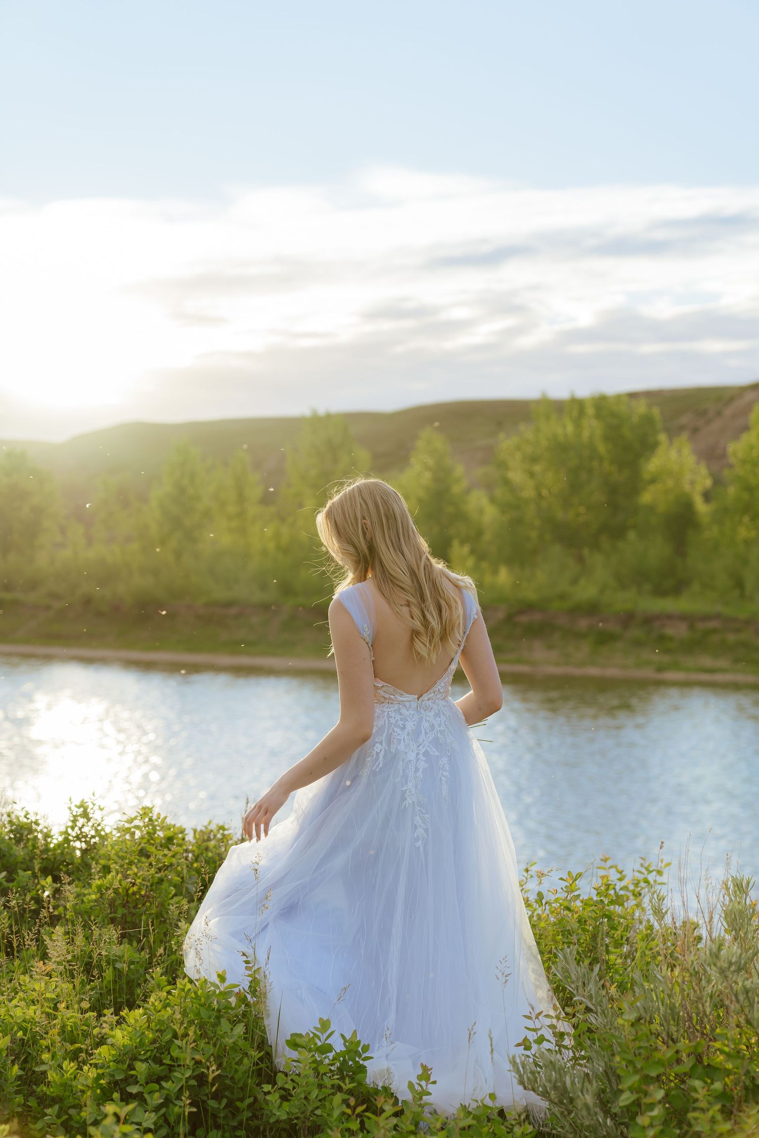 A bride in a white gown stands overlooking a serene lake at sunset.