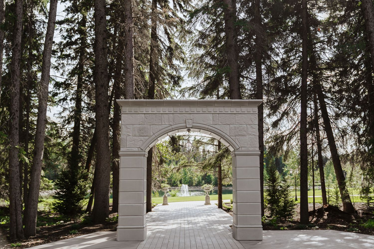 Elegant white archway entrance framed by tall evergreen trees along a brick pathway in a garden setting.