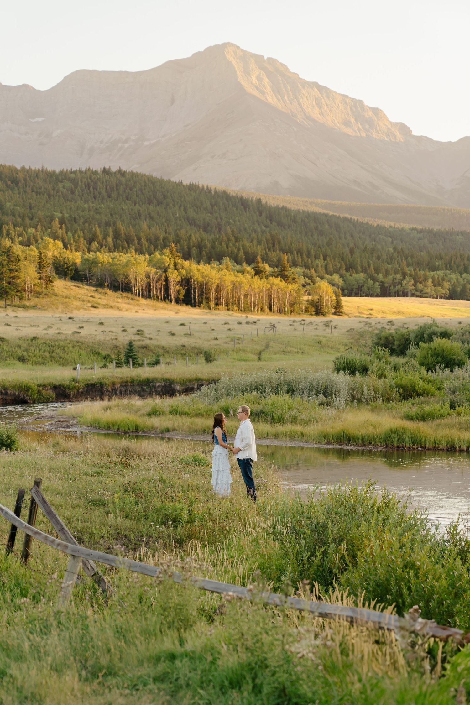 Scenic mountain landscape at sunset with couple standing by pond surrounded by meadows and evergreen forest.