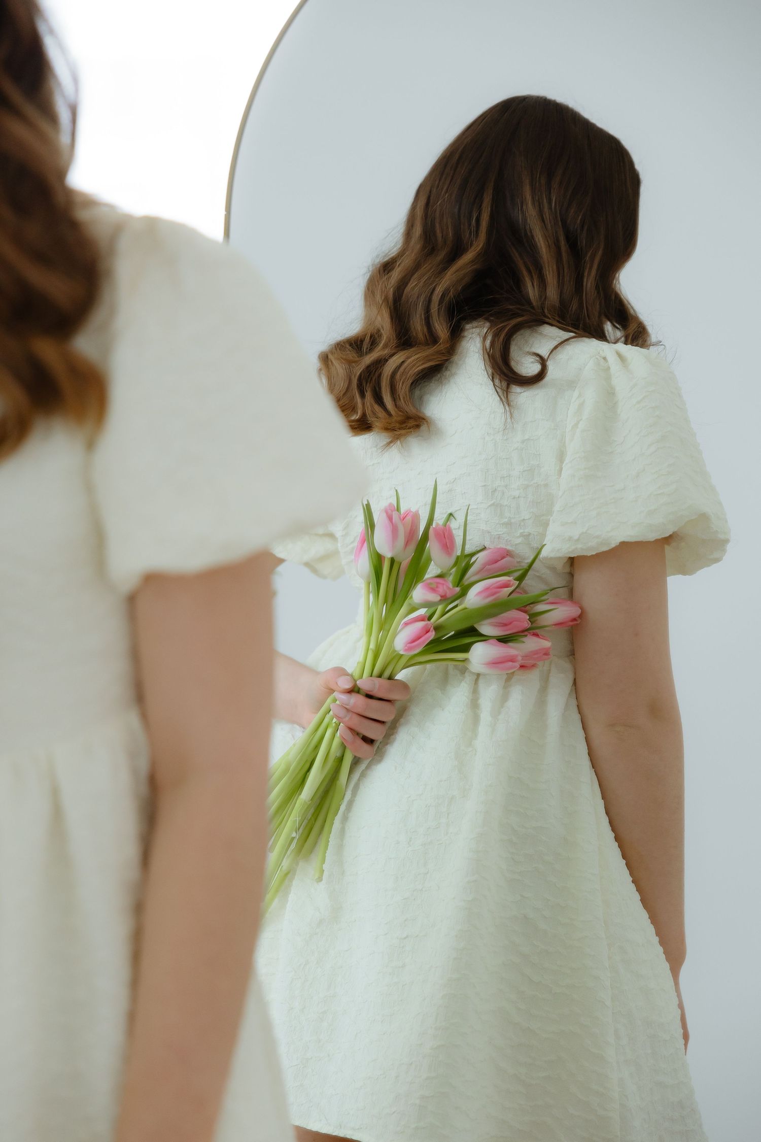 Sara Wiebe in a white dress holds pink tulips behind their back while looking in mirror, creating romantic spring aesthetic.