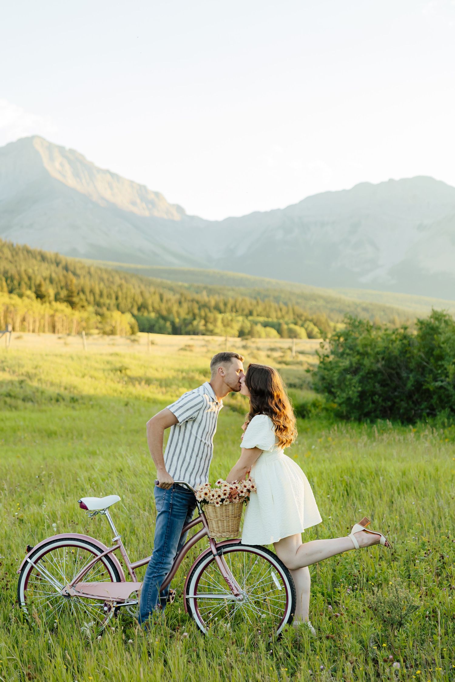 A couple sits together on a vintage bicycle in a scenic mountain meadow at golden hour.