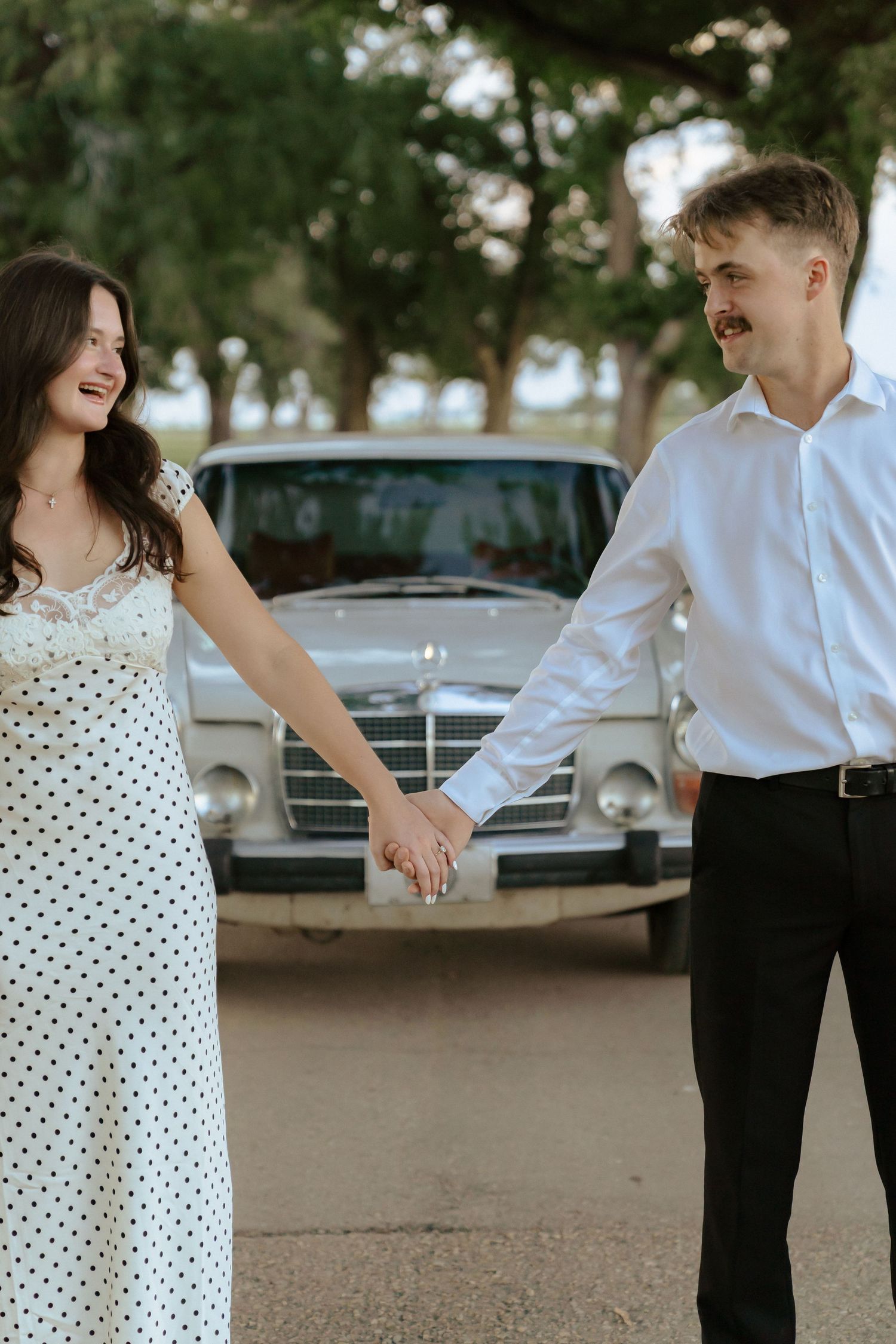 A couple in formal attire shares a playful moment outdoors, with the woman wearing a polka dot dress and man in white shirt.