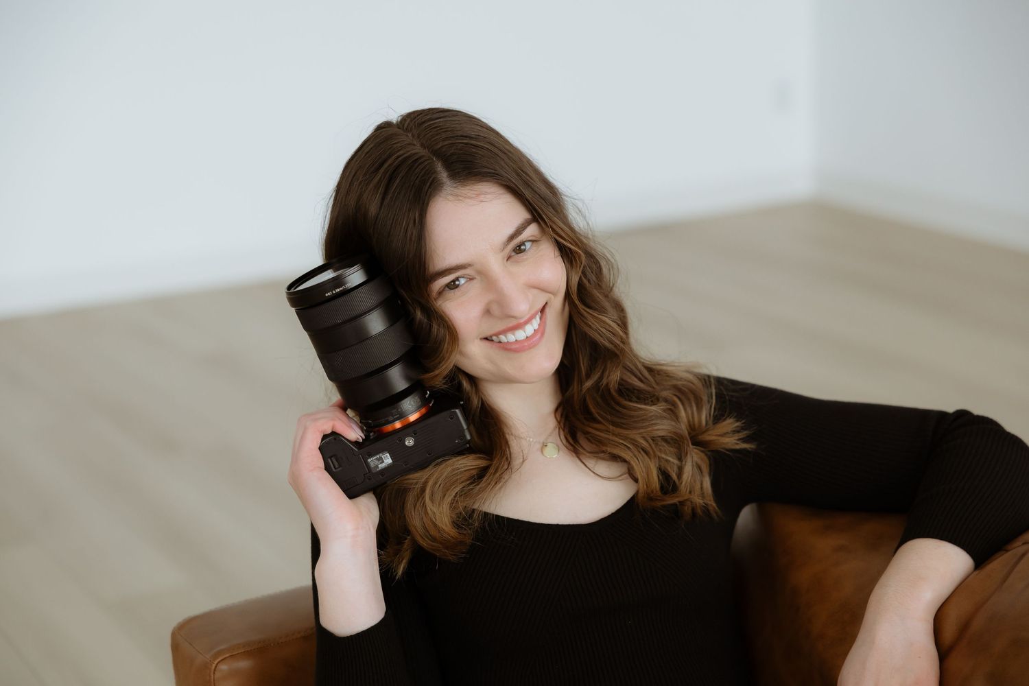 Sara Wiebe a photographer in black attire holds a professional camera lens while sitting and smiling.