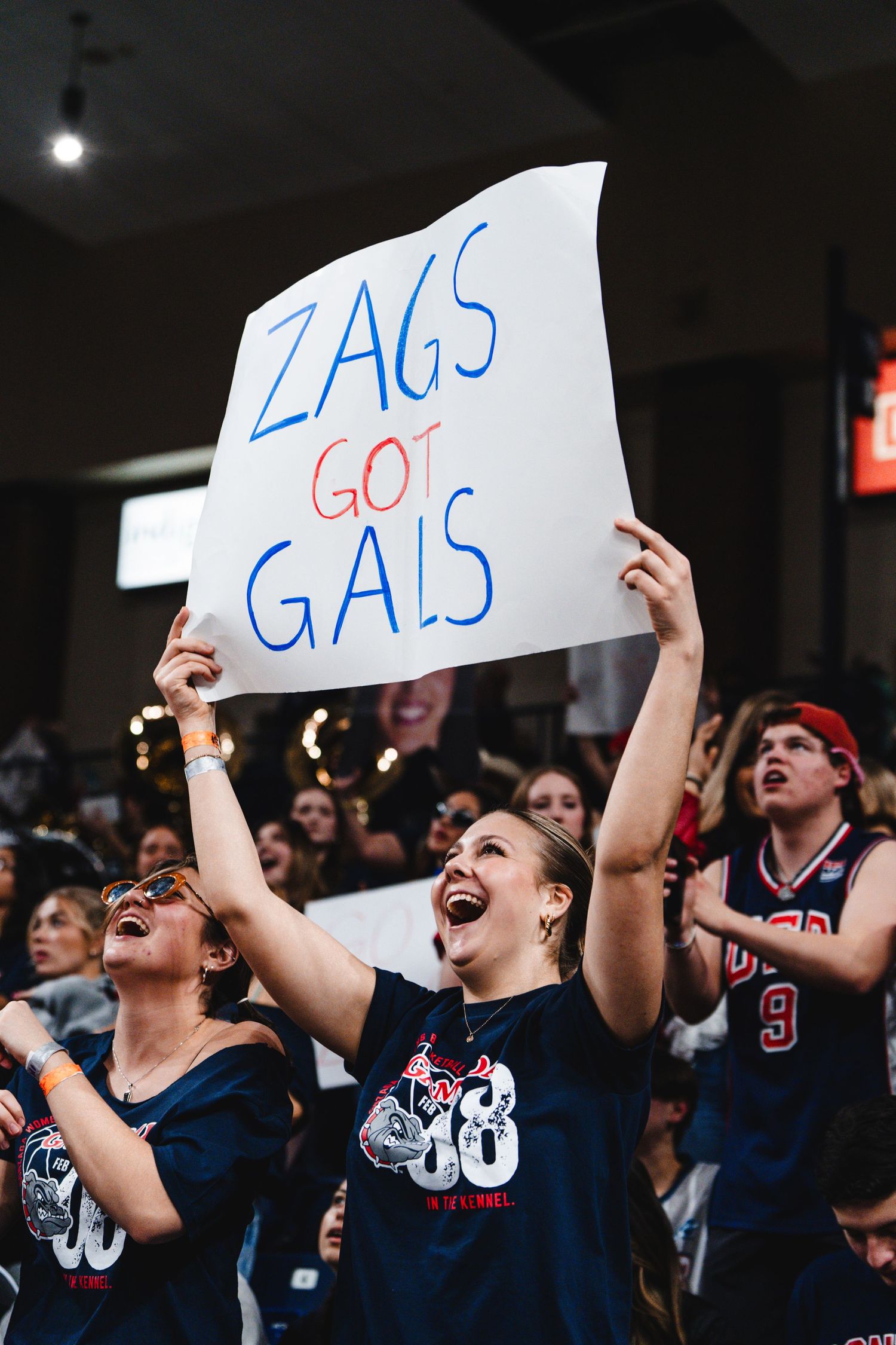 Excited basketball fans in navy jerseys cheer at a game while holding up a 'ZAGS GOT GALS' sign in the crowd.