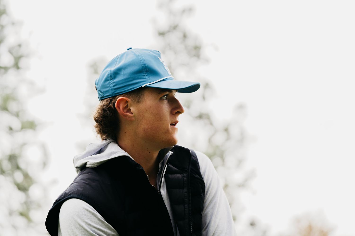 Person wearing a blue baseball cap and black vest looking to the side against a blurred outdoor background.