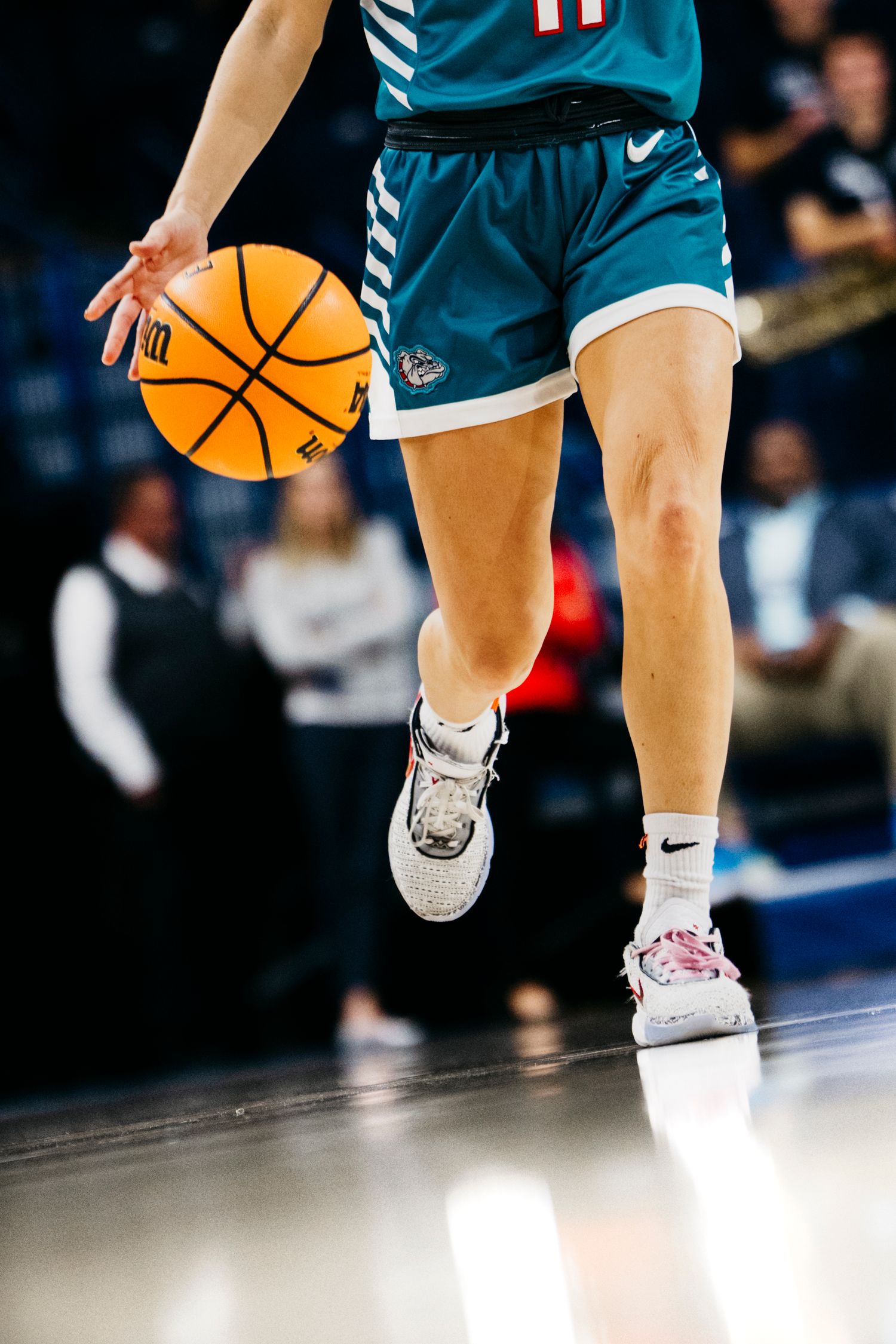 Close-up action shot of basketball player dribbling in teal uniform and Nike sneakers on shiny court floor.