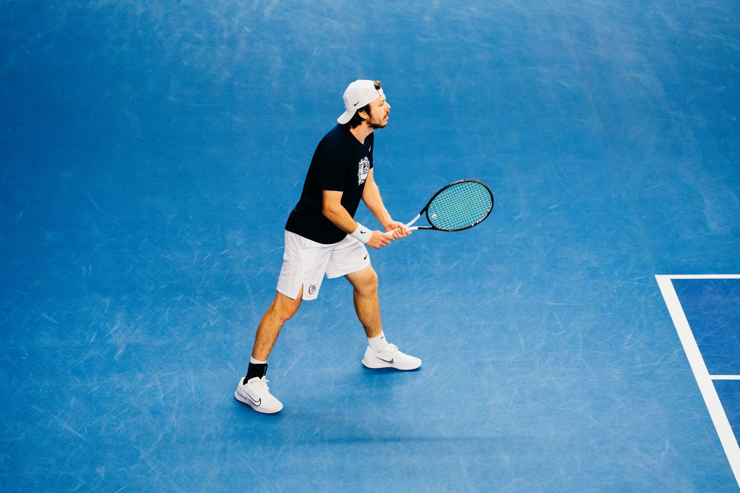 Tennis player in white shorts and black shirt prepares to return a shot on blue court during a match.