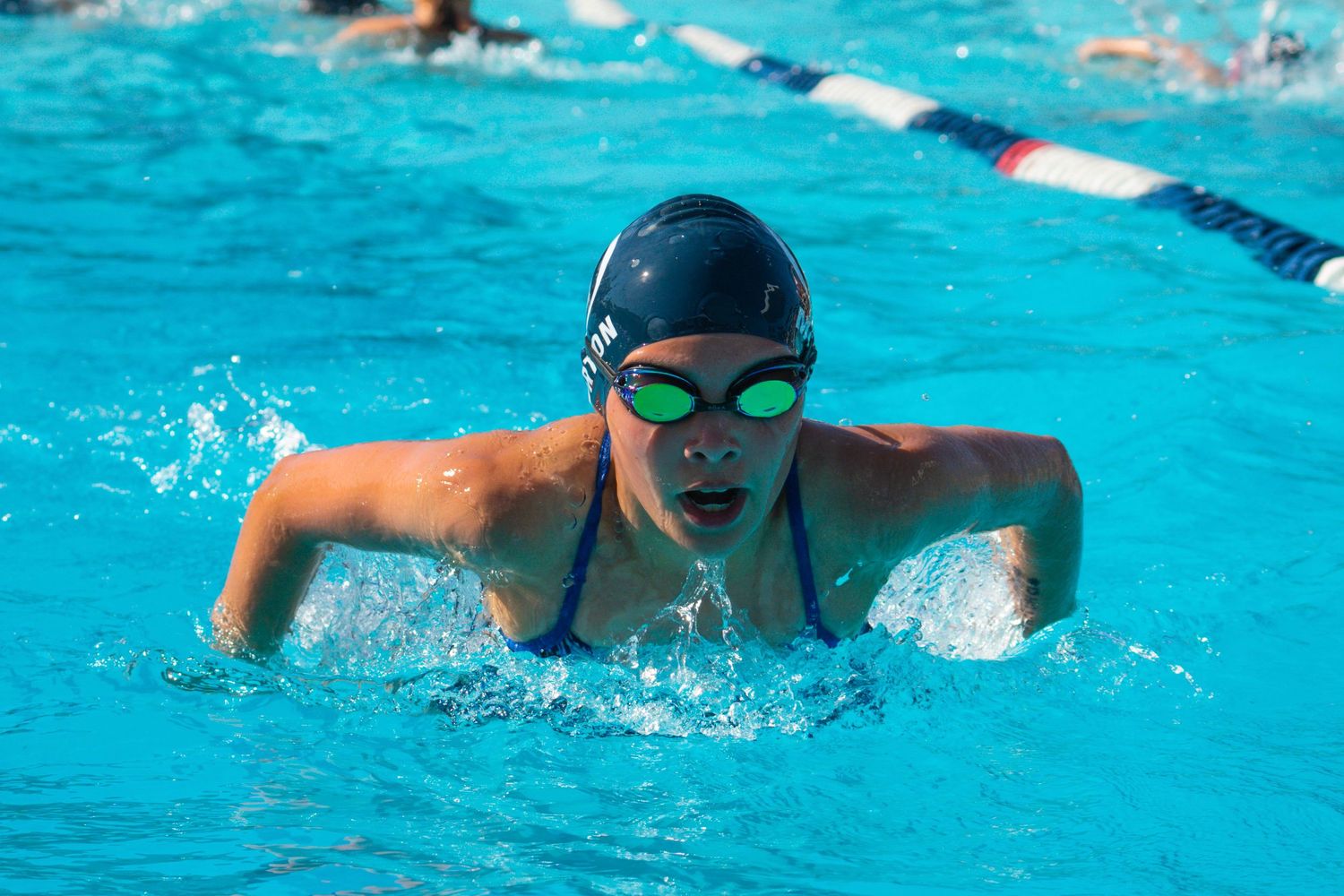 Swimmer performing butterfly stroke in bright blue pool wearing black cap and green goggles during competition.