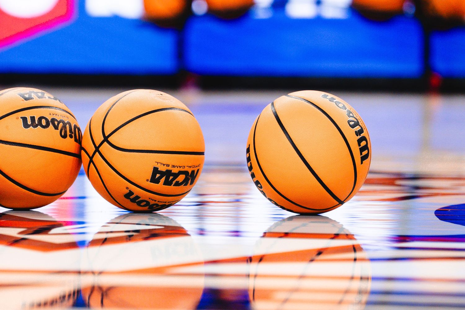 Multiple NBA regulation basketballs rest on a shiny wooden basketball court with red and blue lighting visible.