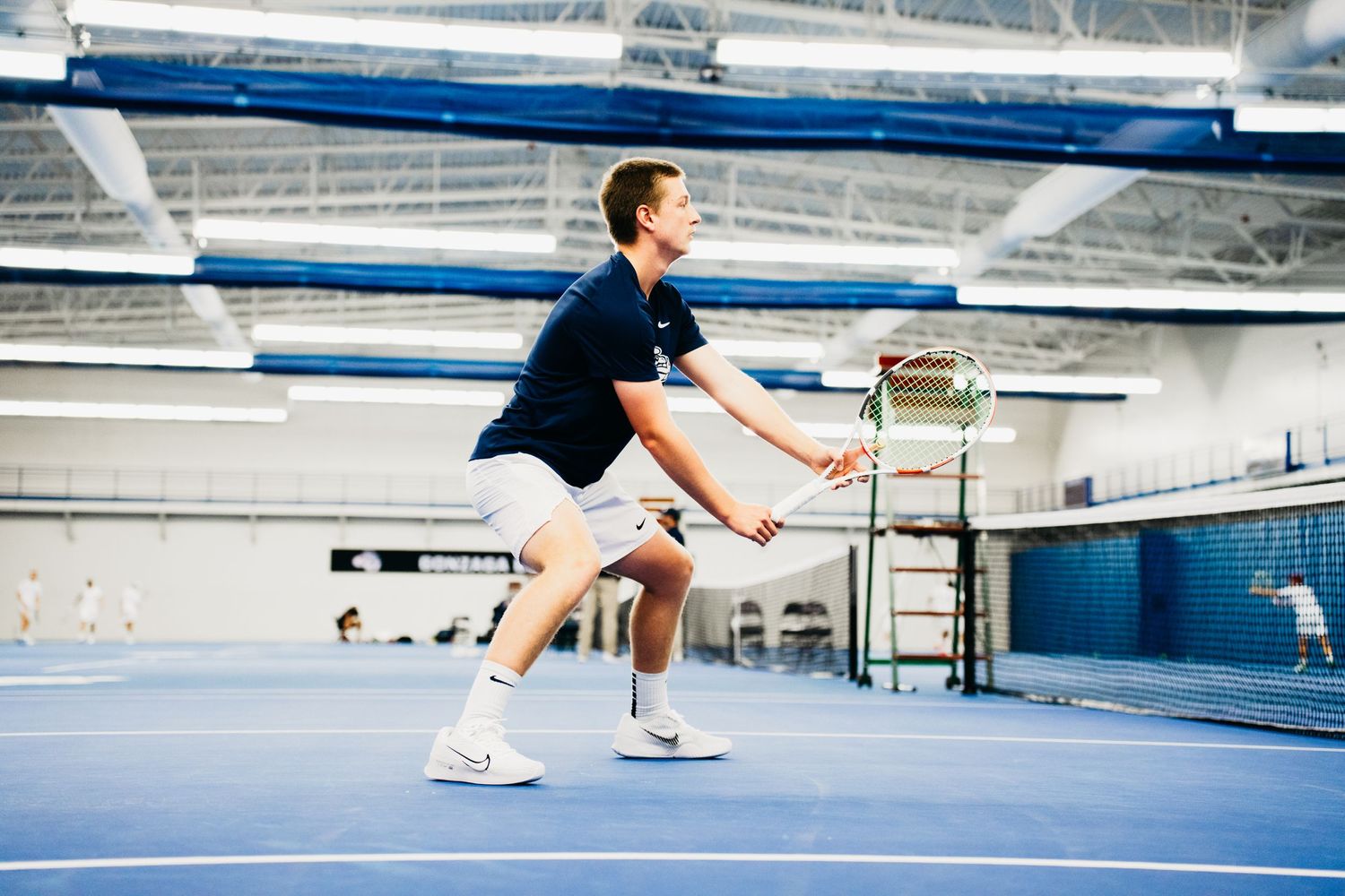 Tennis player in navy blue shirt and white shorts performs a ready position on indoor blue court.