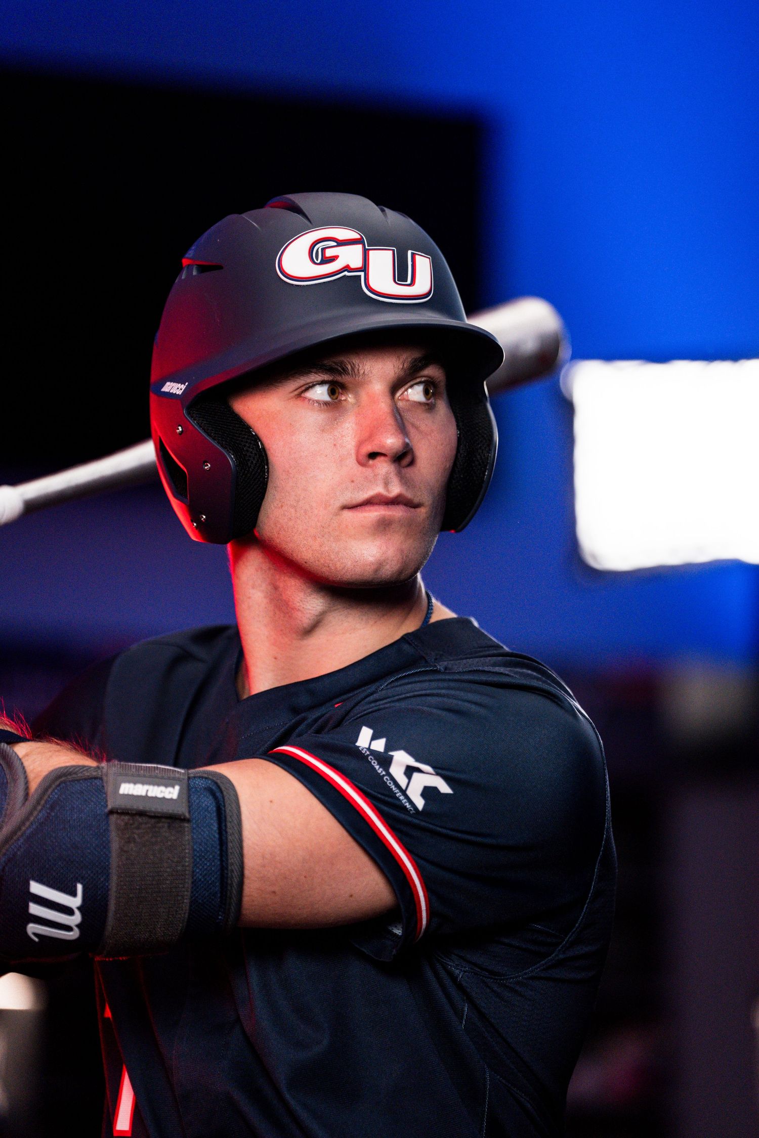 Baseball player in navy uniform and batting helmet with GU logo preparing to bat against blue background.