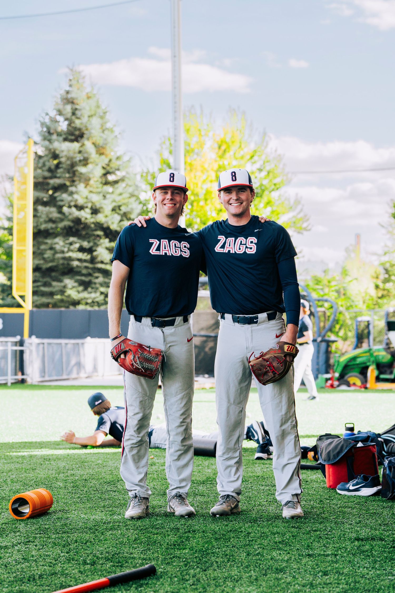 Two Zags baseball players in matching navy uniforms and helmets stand together on artificial turf field during practice.